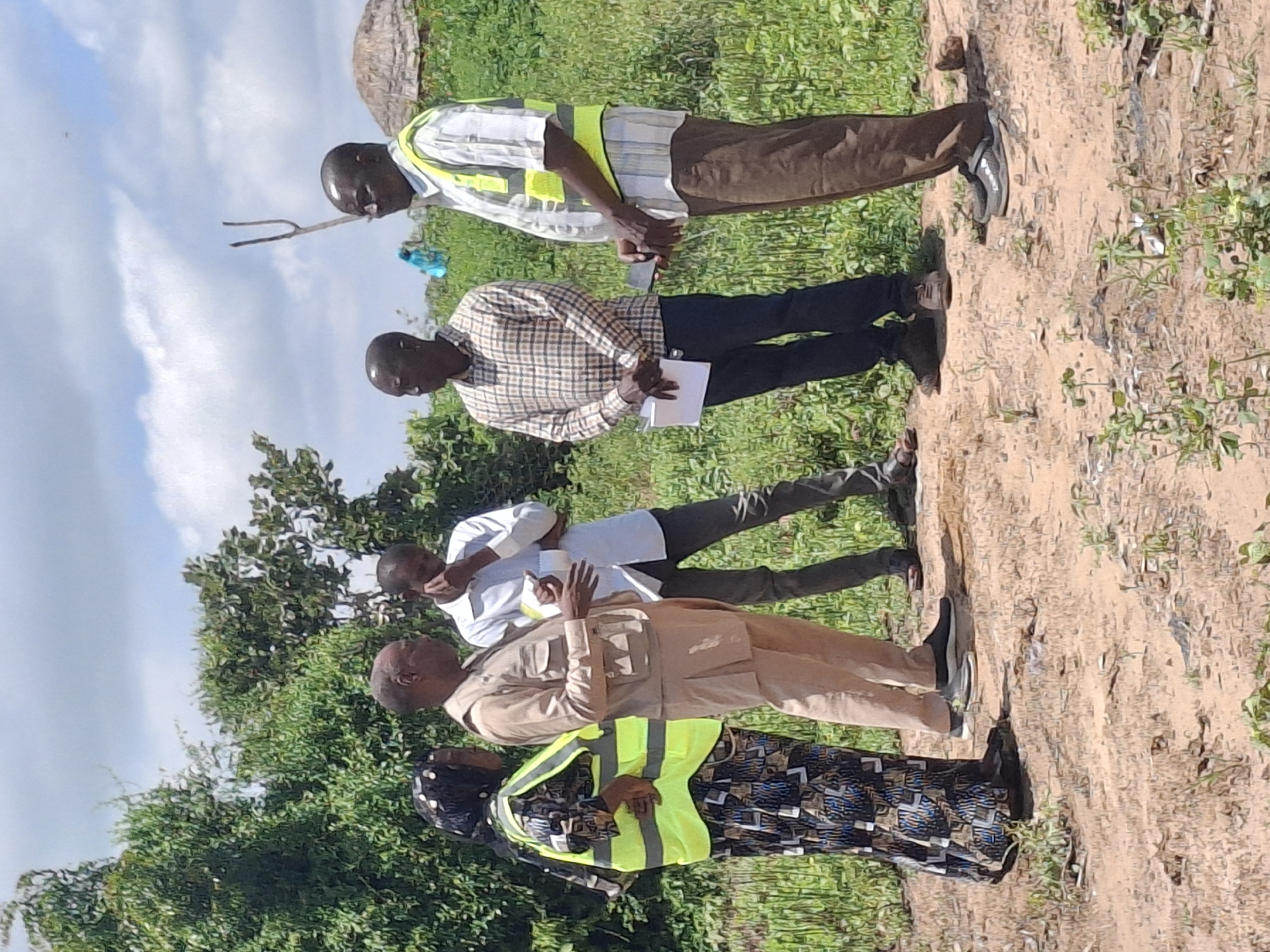 In Lembi, Nigeria, five individuals are gathered outdoors in a rural setting. The ground is dry and dusty, with sparse green vegetation. In the background, there are trees and a thatched-roof structure. The sky is cloudy with patches of blue.

Four men and one woman are present. The woman, on the left, is wearing a headscarf and a patterned dress with a bright yellow safety vest. She is looking towards the men.

The man next to her, wearing a light brown tunic and trousers, appears to be speaking. He has a formal posture.

To his right, a man in a white long-sleeved shirt and dark trousers is seen with his hand near his face, as if contemplating or listening intently. He is holding a small white object.

Further to the right, a man in a checkered shirt and dark trousers is standing with a paper in his hands.

The man on the far right is wearing a white and blue striped shirt with a yellow safety vest and brown trousers. He is holding a dark object, possibly a phone or a small device, and is looking downwards.

The group appears to be engaged in a discussion or a meeting in an open, undeveloped area. The presence of safety vests suggests a possible work-related context.