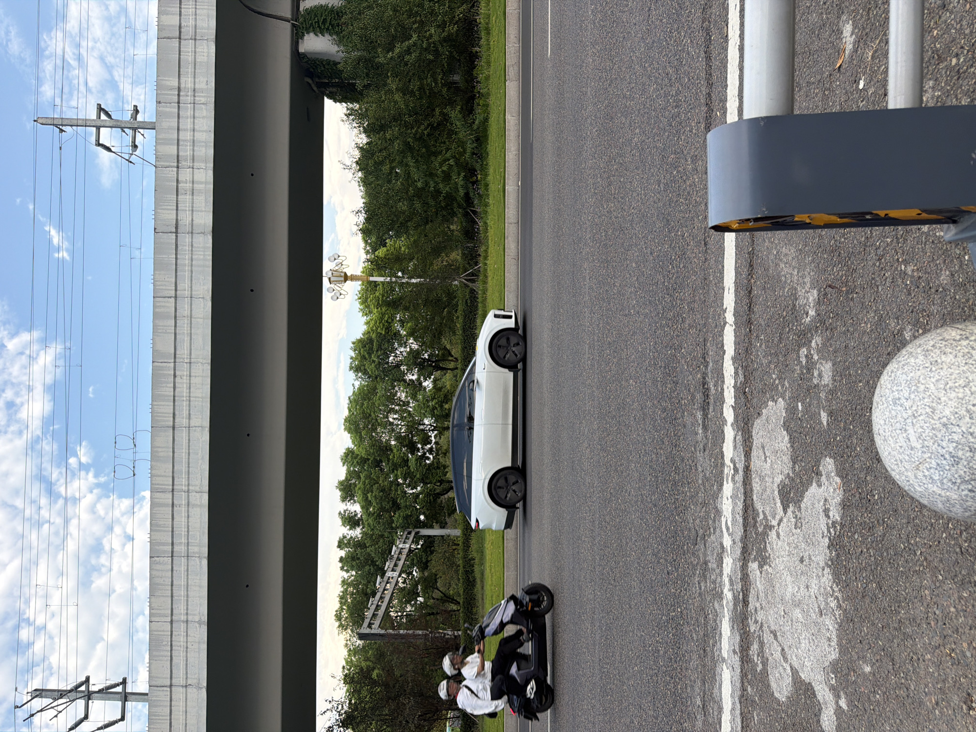The image depicts a street scene under a concrete overpass, likely in Jiangjin, China, given the context provided. Two individuals, wearing white tops and helmets, ride a dark-colored electric scooter, heading to the right. Beside them, a white SUV with a distinctive streamlined design also travels to the right. The road is asphalt, with a white lane marker. To the right of the road, a grassy area is bordered by dense green trees. An ornate lamppost stands among the trees. In the background, a portion of the overpass is visible, with power lines and utility poles extending above it against a sky with scattered clouds. The foreground shows a section of the road with a worn white marking and a textured grey bollard.
