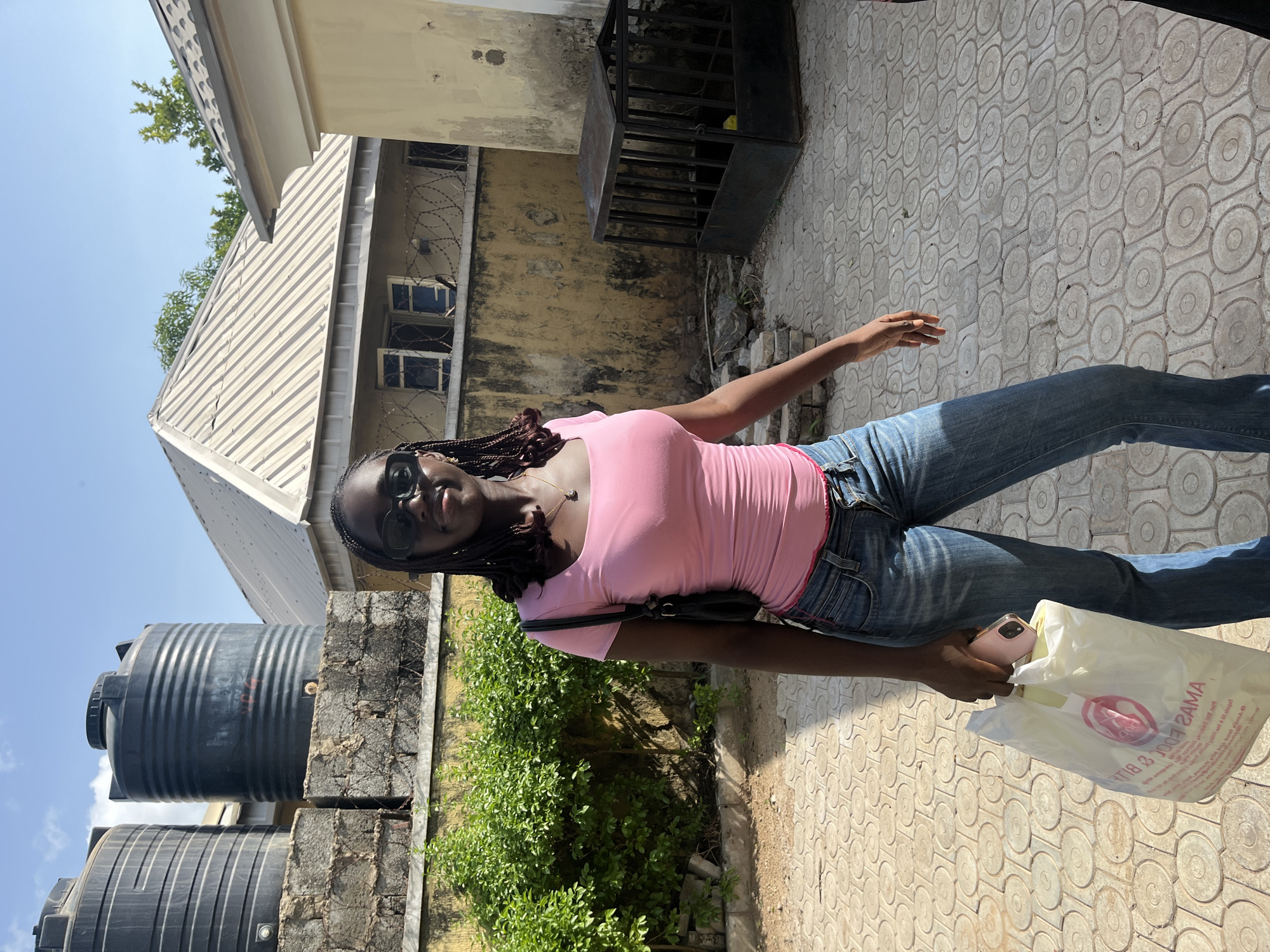 A young woman stands on a patterned concrete pathway, possibly in Abuja, Nigeria, as suggested by the environment. She is wearing sunglasses, a pink top, and jeans, and she carries a plastic bag in her right hand. Behind her are buildings, including one with a corrugated metal roof and windows protected by barbed wire. There are also water tanks and green bushes nearby.