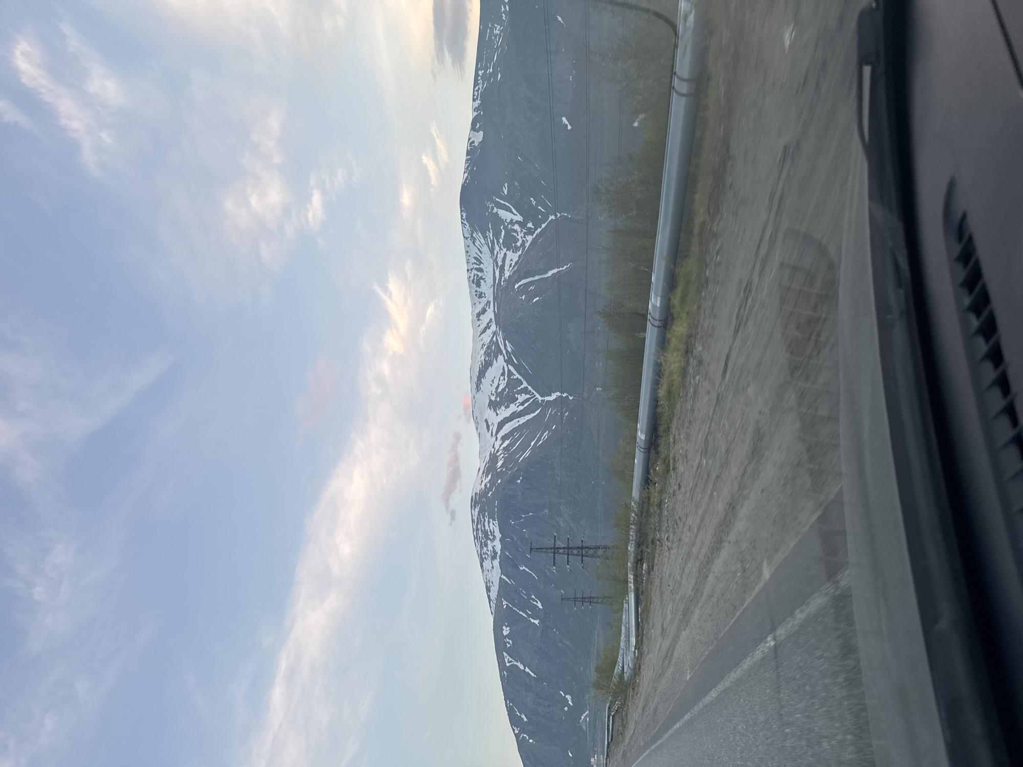 The image depicts a vast, rugged landscape from the interior perspective of a vehicle, looking forward through the windshield. The upper third of the frame shows a pale blue sky with scattered white cirrus and stratocumulus clouds, some exhibiting faint pink and orange hues indicative of early morning or late evening light. Dominating the middle ground are dark grey mountains, characterized by steep slopes and numerous streaks and patches of residual white snow, particularly in valleys and on higher elevations. A small, distinct pink cloud hovers just above a mountain peak in the central background. Below the mountains and extending across the mid-ground, a calm body of water is visible, bordered by sparse green vegetation. Two prominent, multi-legged metallic power line towers are positioned in the mid-ground, to the left of center, with visible transmission lines. In the immediate foreground, a paved road with faint white markings occupies the lower-left, transitioning to an unpaved, rough dirt shoulder on the right. Parallel to the road and the unpaved area, several large, light grey industrial pipes run horizontally across the mid-foreground. Sparse, low-lying green vegetation, including small trees and bushes, grows along the roadside and near the pipes. The dark dashboard and the lower edge of the vehicle's windshield, showing minor reflections, are visible at the very bottom of the frame. No human figures or animals are present. The mountainous terrain with persistent snow, industrial infrastructure, and the specific lighting conditions are consistent with the geographical and industrial characteristics of Kirovsk, located within the Khibiny Mountains in Murmansk Oblast, Russia.