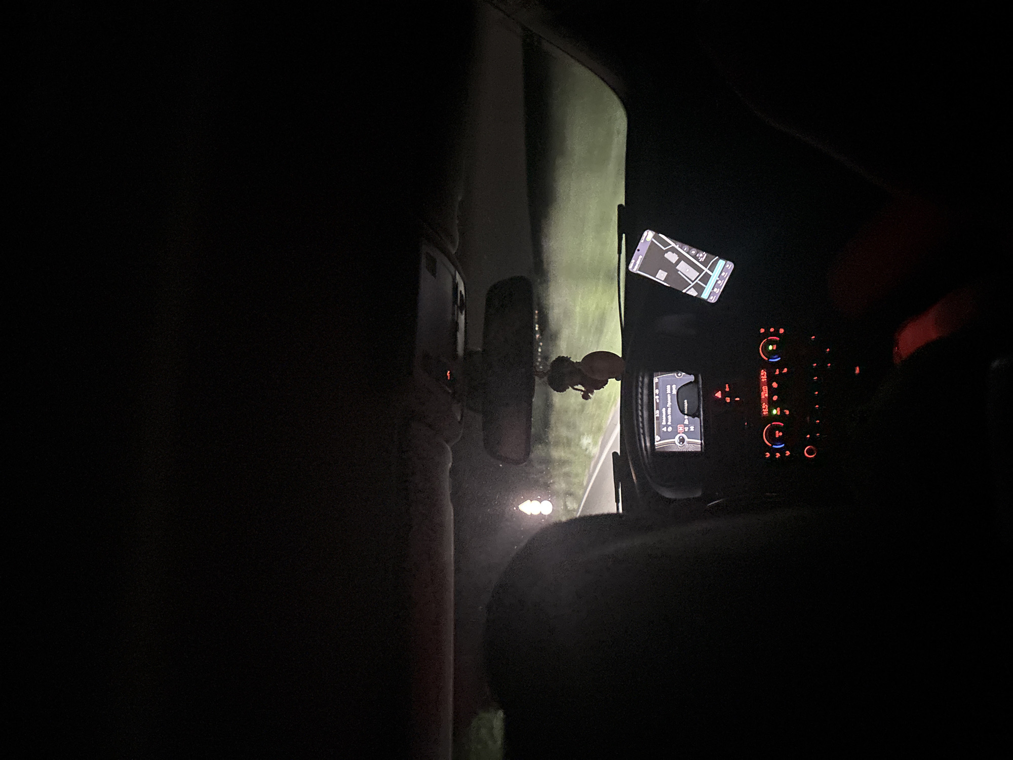 The image depicts the interior of a vehicle at night, presumably in transit. The dashboard is visible, illuminated with various instrument panel lights and a central display showing navigational information. A mobile phone is mounted above, displaying a map. The scene is dark, but the headlights illuminate the road ahead. Through the windshield, a glimpse of the roadside is visible, suggesting a rural or less populated area. The location of Hrabarsko, Bulgaria, cannot be confirmed due to insufficient visual clues within the image. There are no visible persons.