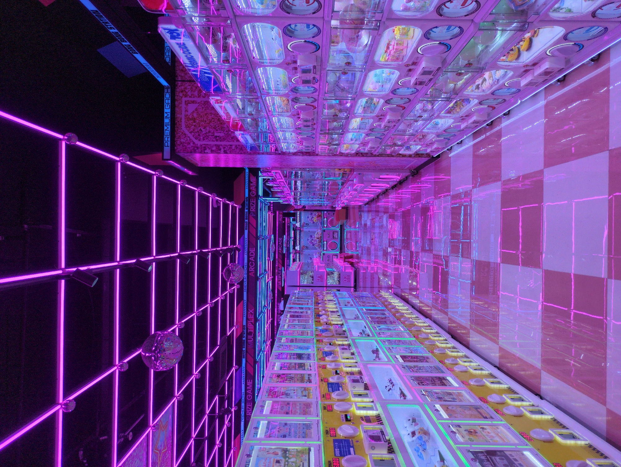 This image depicts the interior of an arcade in Daejeon, South Korea. The perspective is distorted to make the arcade appear longer, more like a tunnel. The walls are lined with prize games and gumball machines illuminated with vibrant pink and blue neon lights. On the left, there is a grid-like structure with neon pink tubes and reflective disco balls attached. The floor and ceiling are reflective. Text such as "PREMIUM GACHA", "BONG BONG STATION", "PRIZE GAME", "MULTIPLEX", and "ARCADE" are visible along the walls.