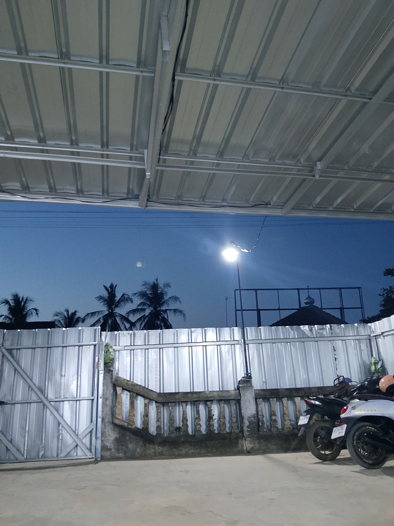 This is an outdoor, medium-shot, eye-level photograph taken at dusk in Negeri Agung, Indonesia. The scene is under a corrugated metal roof supported by a metal frame. The sky is a deep blue, with a street light on the left casting a bright glow, and a faint circular light, possibly the moon, visible higher up. In the background, silhouetted palm trees are visible against the sky, along with a structure that resembles a dome. A corrugated metal fence runs across the middle ground, with a concrete barrier topped with a decorative, aged railing in front of it. On the right side of the image, parked next to the fence, are two motorcycles. The ground in the foreground is a paved concrete surface. The overall atmosphere is quiet and appears to be a residential or communal outdoor space at the end of the day.