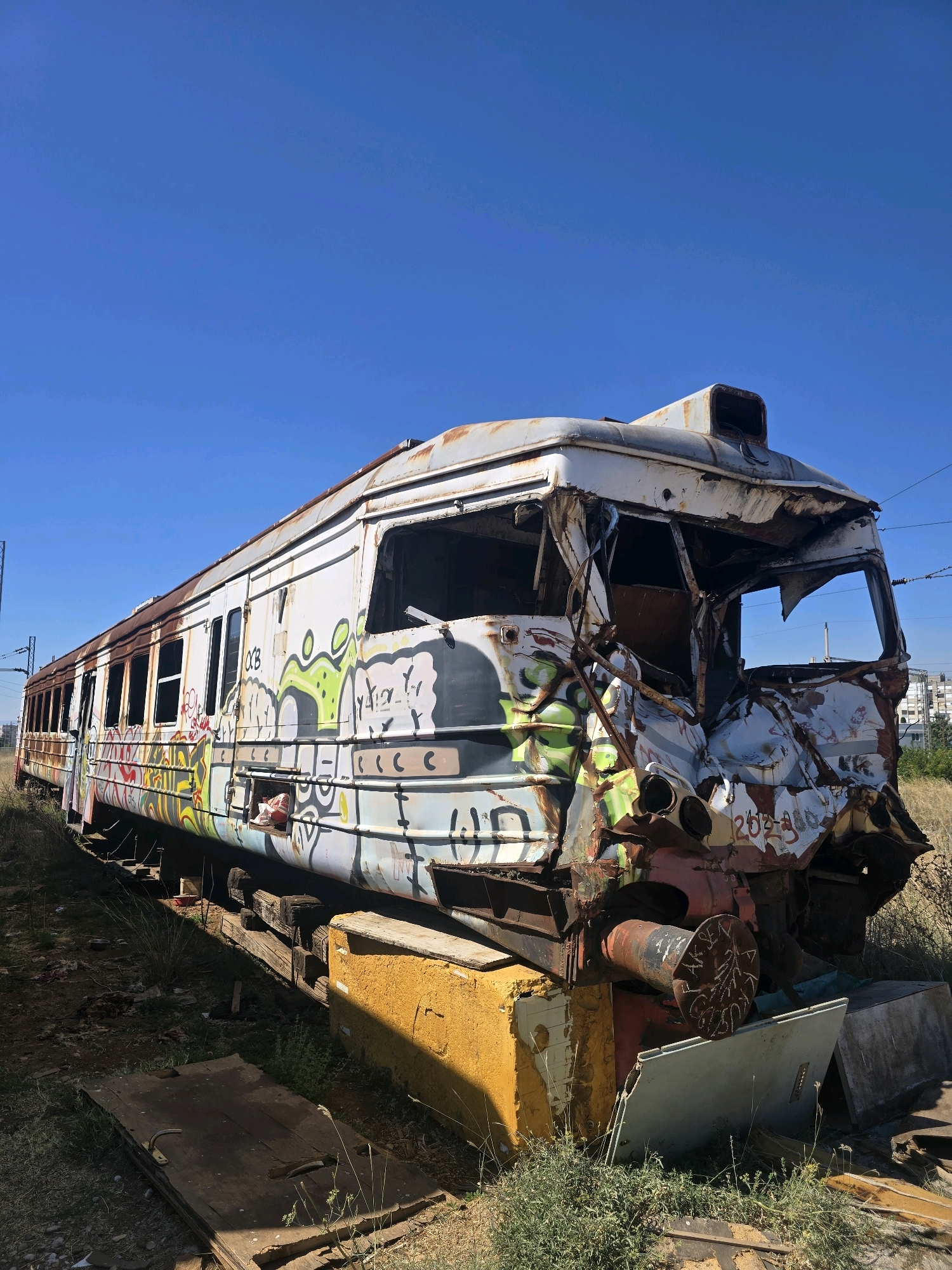 A damaged, graffiti-covered train car rests on tracks outdoors under a clear blue sky. The train car is white with extensive rust and colorful graffiti. The front of the train car is severely damaged and crumpled. The surrounding environment appears to be an overgrown, undeveloped area with dry grass and weeds. There are some wooden beams and debris on the ground near the train. In the background, a few buildings are visible, suggesting an urban or suburban setting. The image was captured in Podgorica, Montenegro, specifically at the abandoned train depot, often referred to as the "old train station" or "Old Railway Station Podgorica."