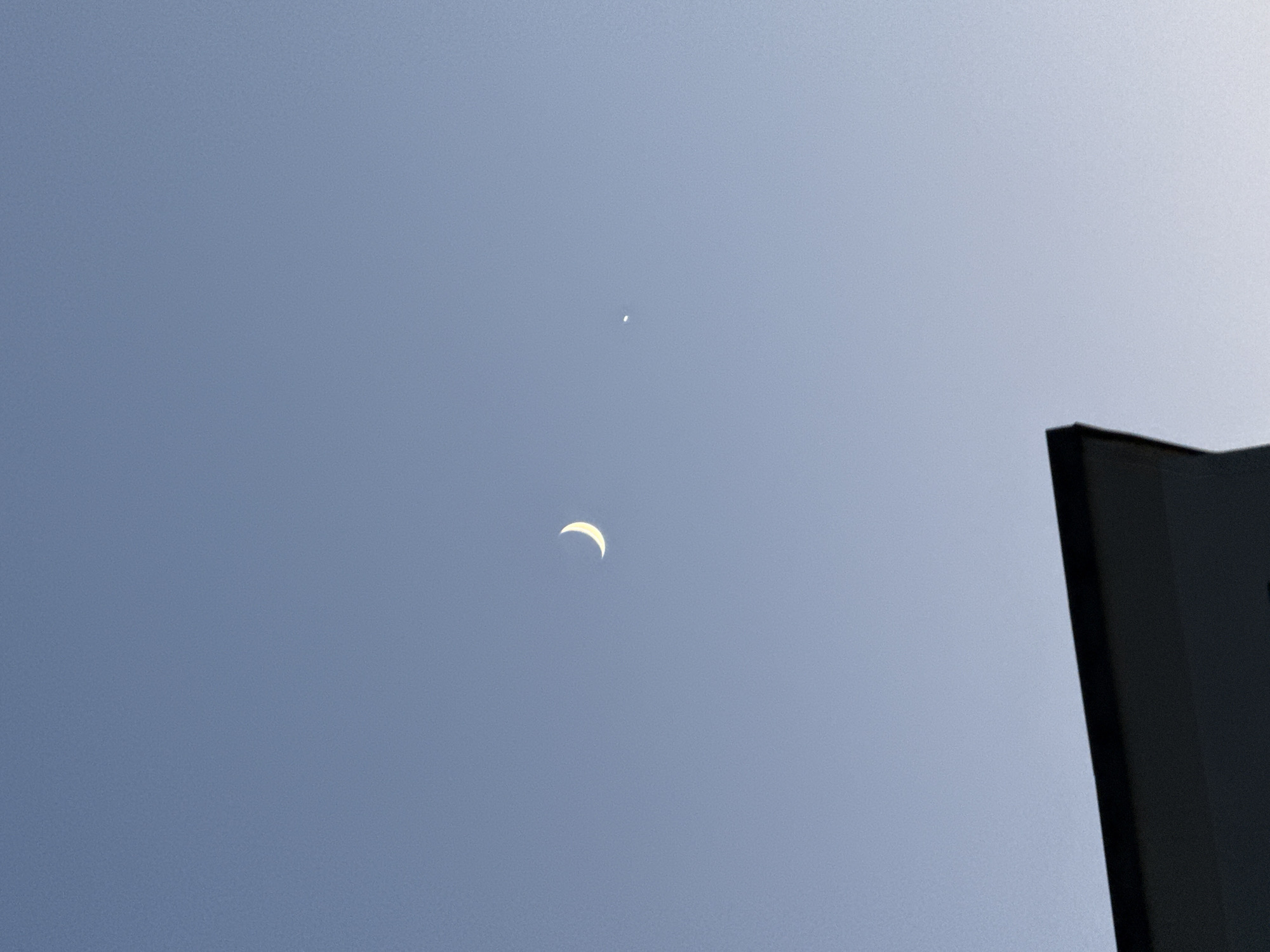 A crescent moon is visible in the clear, light blue sky over Glendale Heights, United States of America. To the right of the moon, a small, bright object, likely a planet, is also visible. In the foreground, the dark roofline of a building cuts across the bottom of the frame. The lighting suggests it is either early morning or late evening.