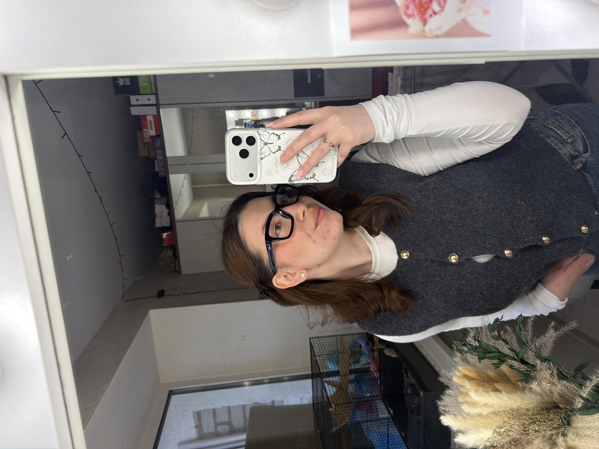 A woman is taking a selfie in a mirror. She has brown hair, glasses, and is wearing a white shirt with a grey cardigan. She is holding a white phone with a butterfly design on the case. The reflection in the mirror shows a room with a window, shelves with boxes, and a cage. There are also flowers visible in the lower right corner. There is no information in the image indicating that the location is Chaikovsky, Russia.