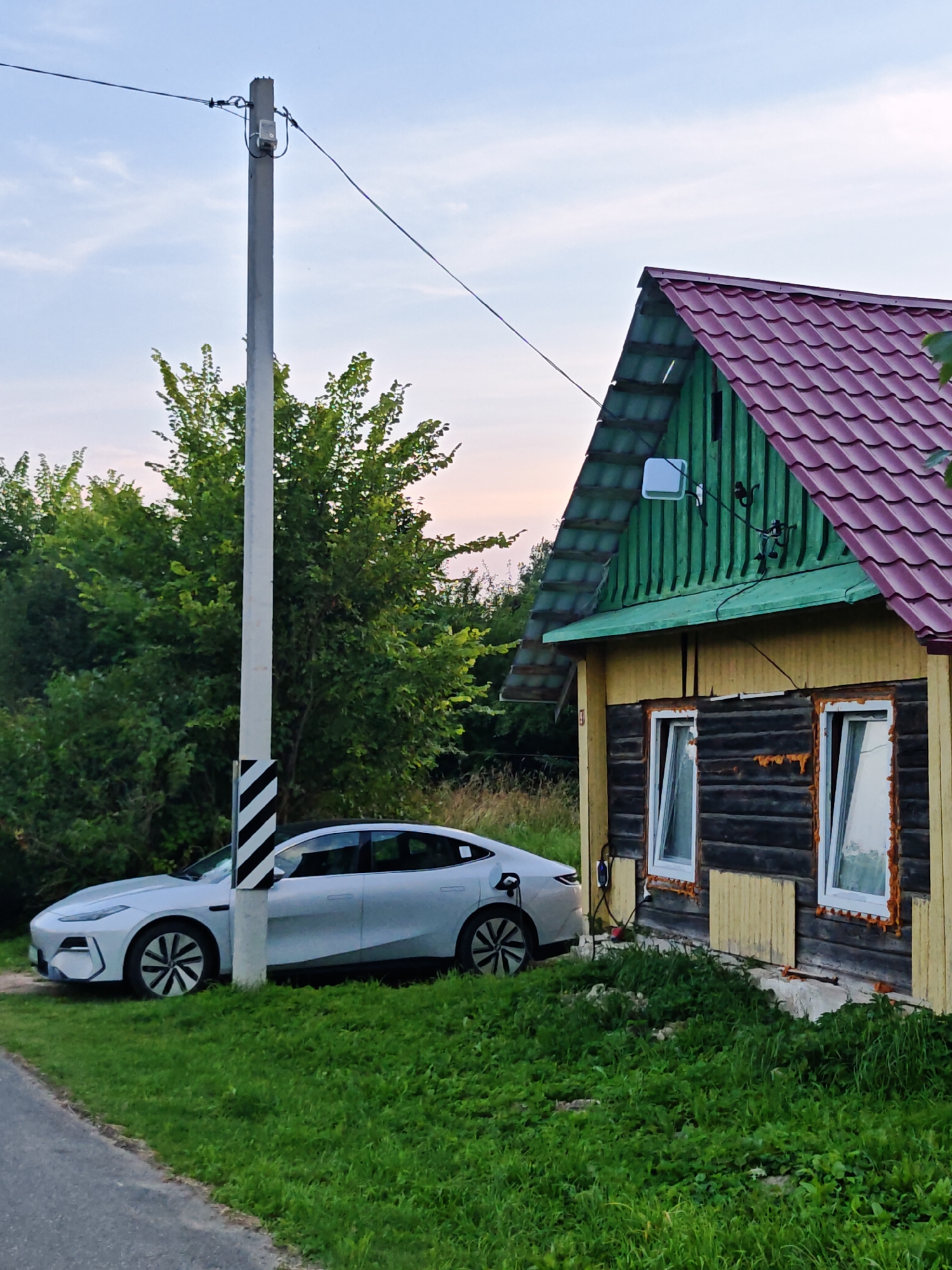 In the city of Valozhynski selski Savet, Belarus, a silver electric car is parked on the grassy shoulder of a road. The car is connected to a charging station located next to a wooden house. The house has a green upper story and a yellow lower story with two visible windows. The roof is made of red tiles. A concrete utility pole with black and white striped markings stands behind the car. Lush green trees and vegetation surround the scene under a pale sky with scattered clouds.