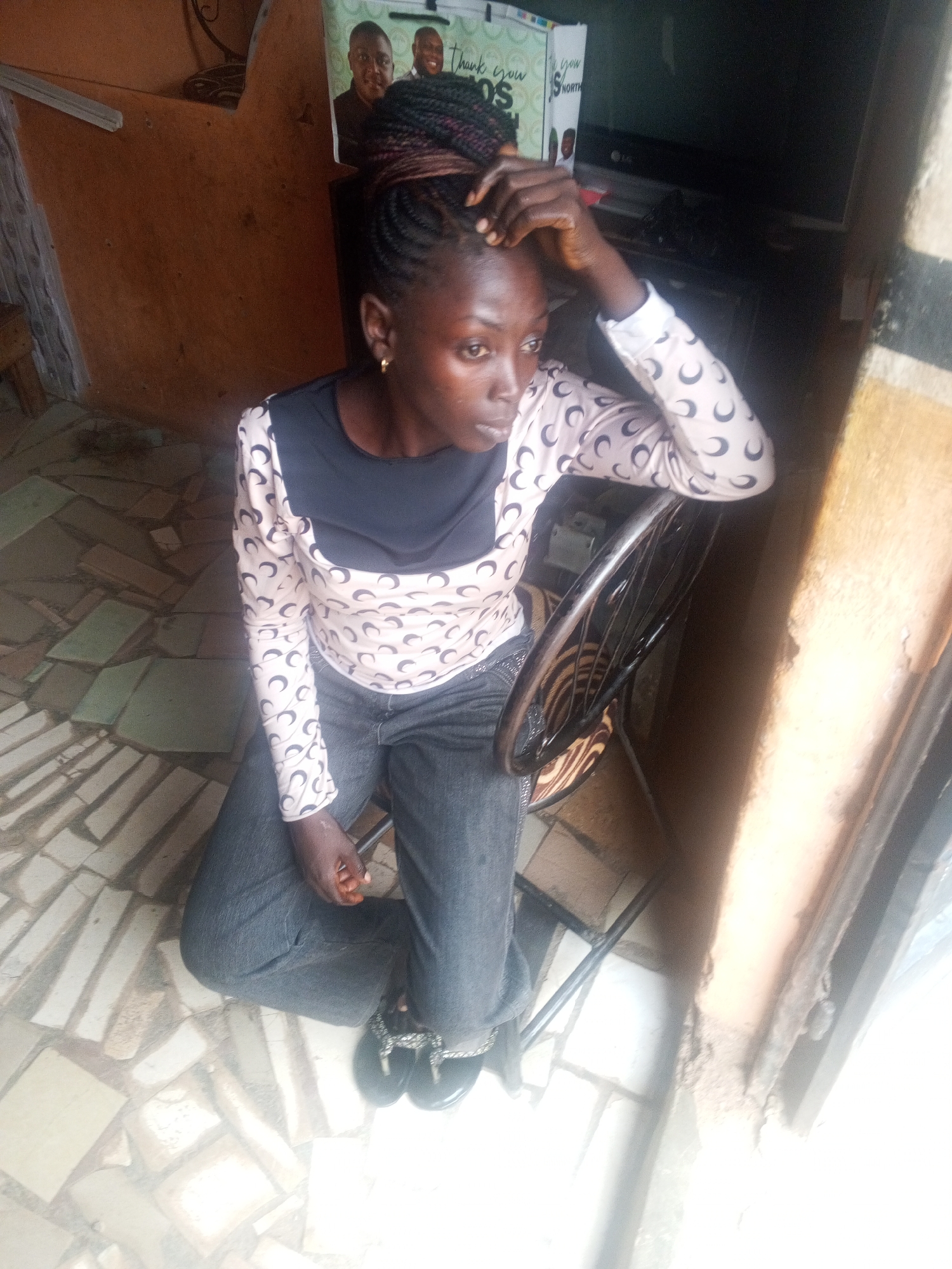 A young African woman is sitting on a chair indoors. She wears a long-sleeved shirt with crescent moon patterns and dark jeans. Her hair is styled in braids. She rests her hand on her head, looking towards a bright window. The floor is tiled in a random rectangular pattern. The location is in Gwong, Nigeria.