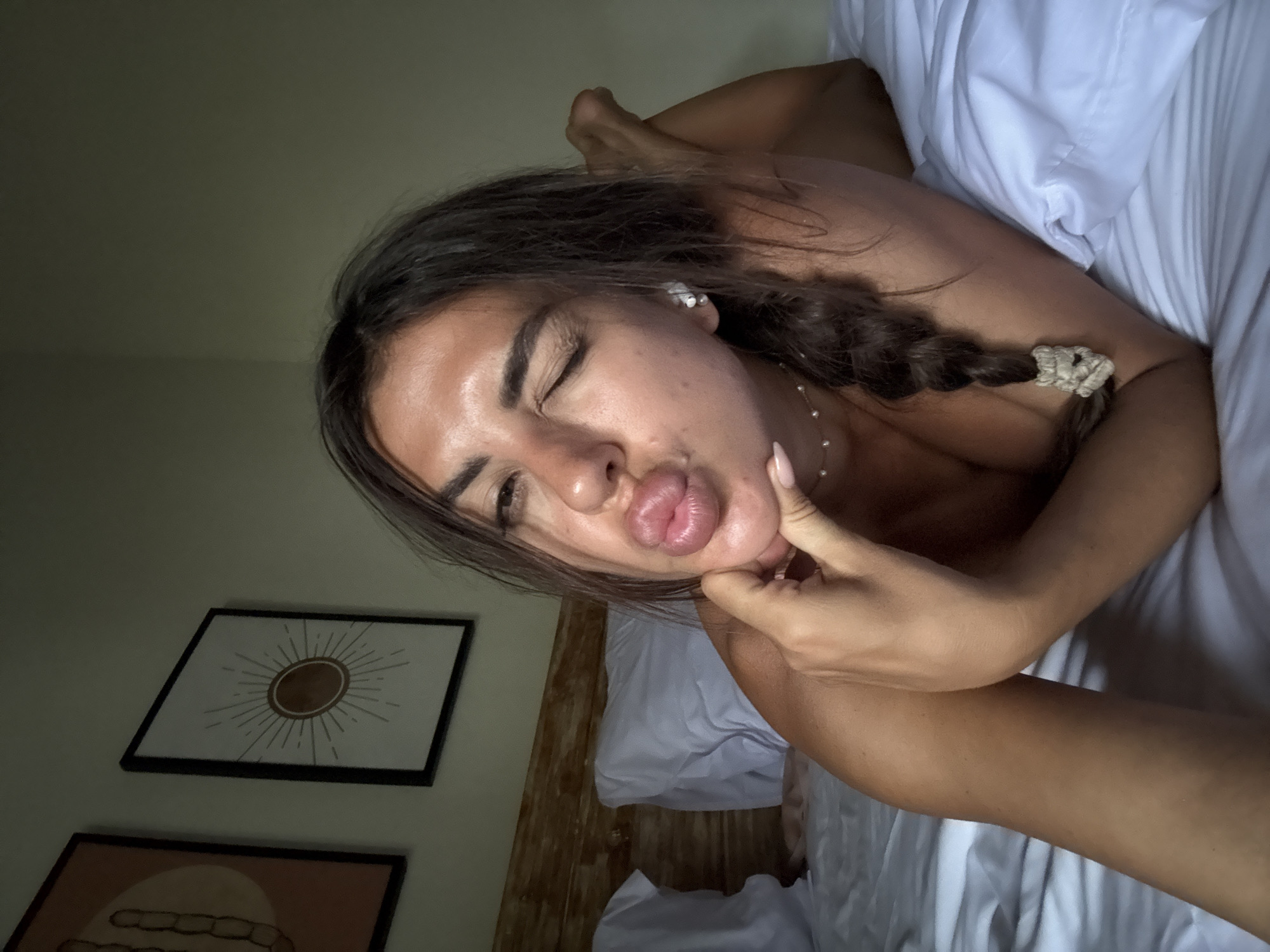 A tanned-skinned woman with dark hair braided on one side and a small pearl necklace is reclining in a bed with white linens. She is wearing a small white earbud in her left ear. Her hand is touching her face, and her lips are puckered. A framed artwork depicting a sun is visible on the wall behind the bed. The setting appears to be a bedroom, likely in a residence or hotel room, based on the artwork and bed linens. The location may be North Kuta, Indonesia.