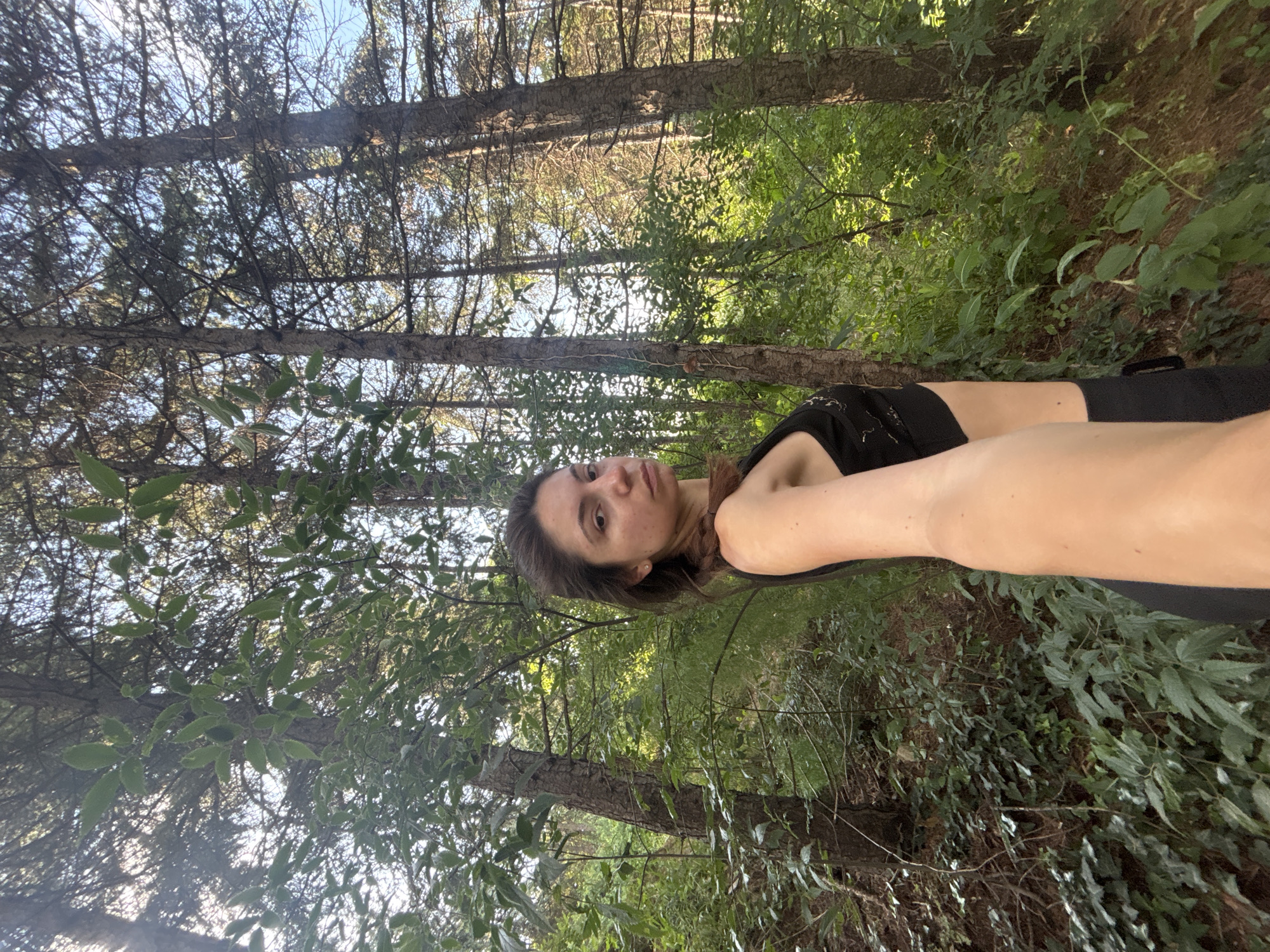 The media file is a selfie taken by a young woman lying down in a forest setting. The woman is in the foreground, with her head and torso visible. She has long, dark brown hair and is wearing a black sports bra. Her eyes are open, and she is looking slightly upwards. Her left arm is extended towards the camera, and her right arm is bent at the elbow.
The background of the image is a dense forest with tall trees and lush green foliage. Sunlight filters through the leaves, creating dappled patterns of light and shadow. The ground is covered with leaves and undergrowth. The overall impression is one of a peaceful and natural environment.
The time of day appears to be daytime, likely late afternoon or early evening, given the angle of the sunlight. The weather seems to be clear and warm, conducive to outdoor activities.
The location is Varna, Bulgaria, as indicated by the context provided. This suggests the woman is in a natural area near the Black Sea coast.
There is no visible text in the image. The mood conveyed is one of relaxation and enjoyment of nature.