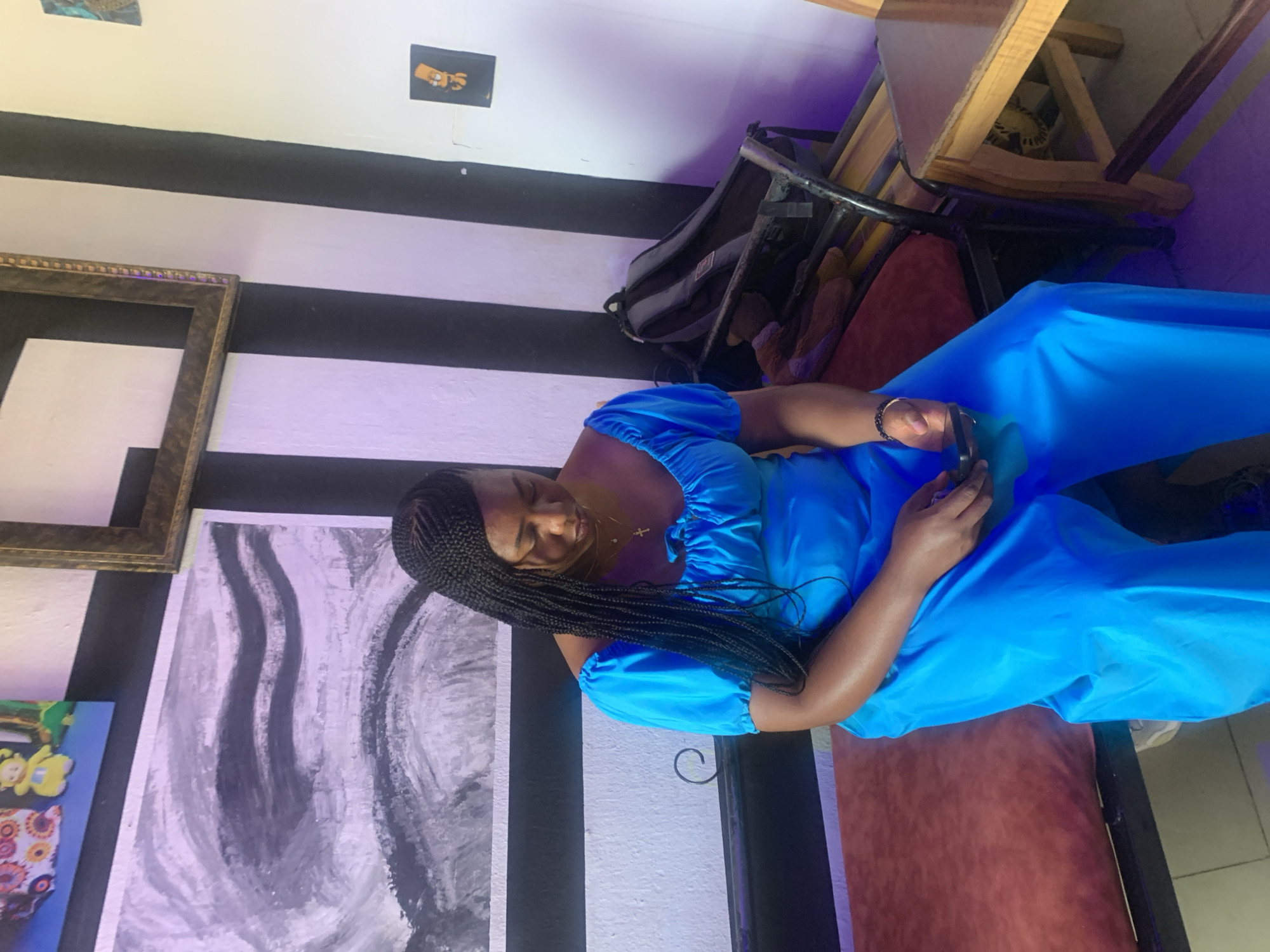 The image captures a young Black woman seated indoors, engrossed in her smartphone. She has long, dark box braids and is wearing a bright blue outfit, which appears to be a jumpsuit with puffed sleeves. A small cross pendant necklace adorns her neck, and she has a silver bracelet on her left wrist. Her posture suggests she is relaxed and focused on her device.
The background features a wall painted with prominent black and white horizontal stripes. Several pieces of artwork are displayed: a large abstract painting with swirling grey, black, and white patterns, a smaller colorful piece with yellow figures and floral designs, and an empty golden ornate picture frame. Higher on the wall, there are smaller framed images; one clearly depicts a character resembling Bart Simpson, with the text "Sips!" visible on a yellow banner next to him.
Behind the woman, parts of a dark-framed chair with a red cushioned seat and a wooden table are visible. A dark grey backpack or bag is stowed beneath the furniture. The lighting in the room has a noticeable purple tint in certain areas, contributing to the ambiance of the space. The setting suggests a casual establishment, possibly a cafe or lounge, given the decor and seating arrangement.