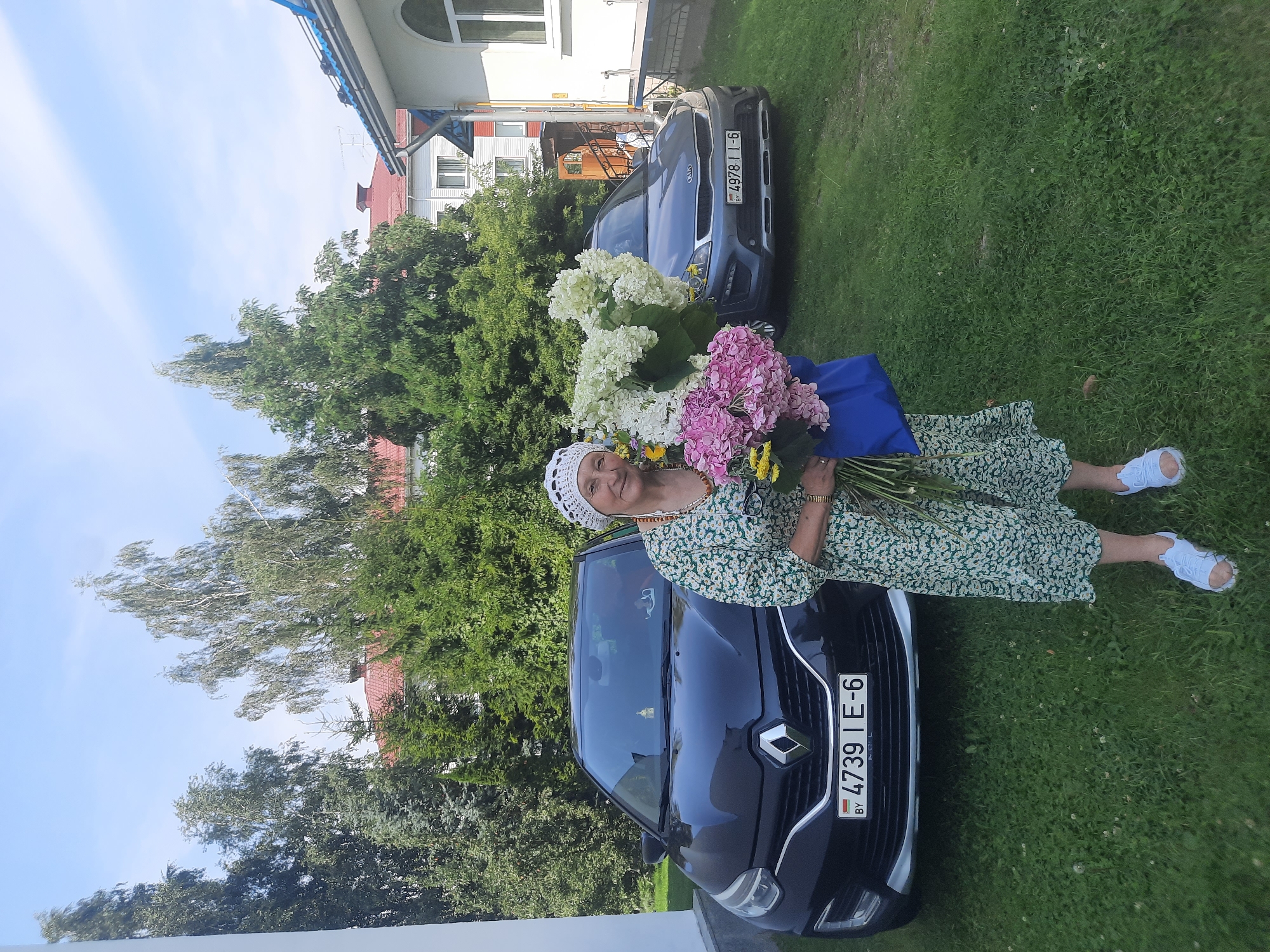 An elderly woman, wearing a floral dress and a white headscarf, stands outdoors holding a large bouquet of pink and white hydrangeas and yellow flowers. She is positioned in front of two cars in a grassy yard with trees and buildings in the background. The black car on the left has a license plate that reads "BY 4739 IE-6". The gray car on the right has a license plate that reads "BY 4978 | I-6". The environment suggests a residential area in the city of Shklov, Belarus, possibly a private yard or driveway.