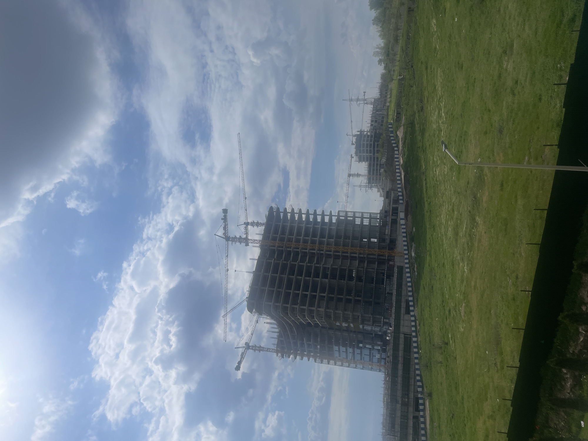 The image depicts a construction site in Yerevan, Armenia, showing unfinished high-rise buildings under construction, surrounded by multiple tower cranes. The buildings have concrete frames, but no facade. To the side of the buildings, there is a large grassy area, partially fenced off. The sky is cloudy.
