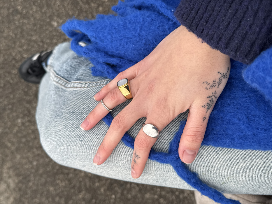 A person's hand adorned with three rings rests on their lap. The rings are on the index, middle, and pinky fingers. A gold ring with a gray stone is on the index finger. A slim ring with small, light-colored stones is on the middle finger. The pinky finger holds a round silver ring. A small black tattoo decorates the top of the hand. The person wears jeans and a blue sweater or scarf. Part of a black shoe is visible in the background.