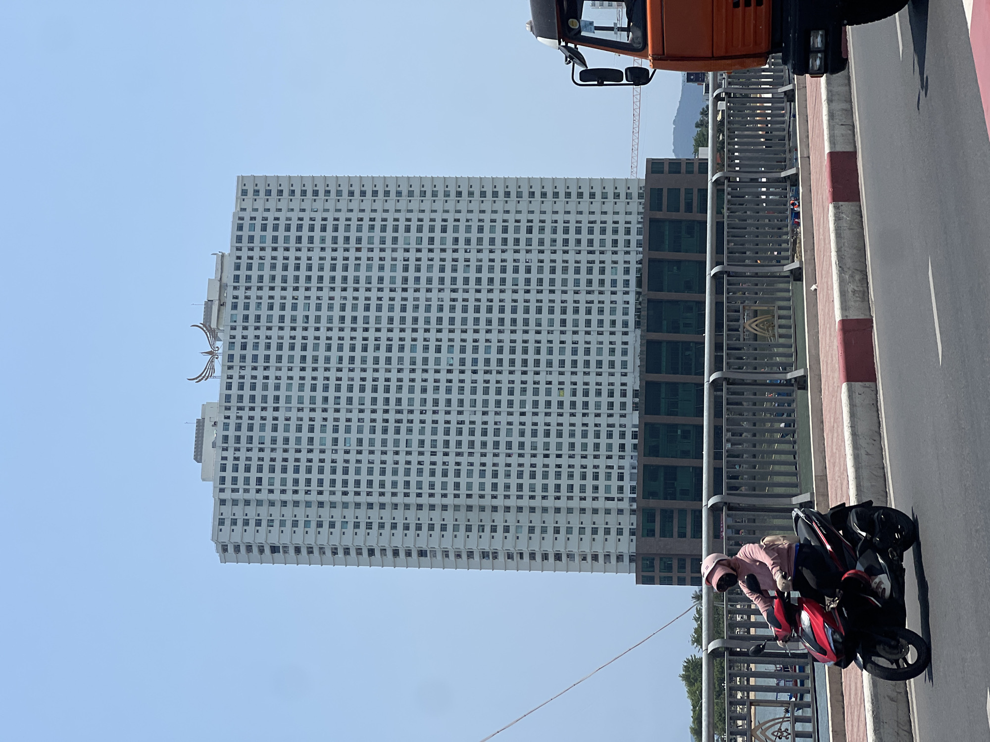 A high-angle, oblique shot depicts a scene in Nha Trang, Vietnam. A tall, rectangular, white building dominates the skyline, densely populated with windows. To the right of the building, a shorter, dark-brown structure can be seen. Adjacent to the right, a gray bridge with a red and white striped border is visible. On the bridge, a person in pink attire is seated on a red and black motorbike. Another motorbike is parked nearby. At the top right of the frame, the cabin of an orange truck is visible. A crane can be seen in the background. The sky is clear and light blue. The scene appears to be captured during daylight.