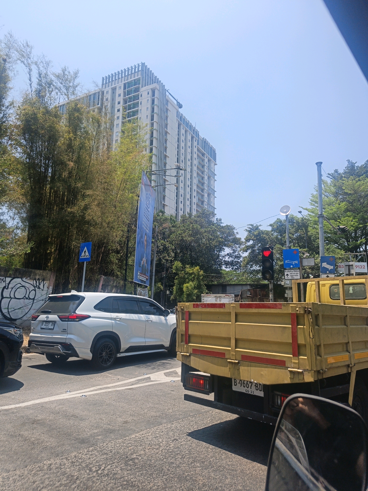 The image depicts a street scene in North Jakarta, Indonesia, on a sunny day. In the foreground, the rear of a yellow flatbed truck is visible, showing its license plate B-9667 BD and the date 05-31. To the left of the truck, a white SUV with license plate B-2217 RH is stopped at a traffic light, which is currently red. Behind the SUV, a large, modern high-rise building is under construction, with bamboo trees lining the street in front of it. Several traffic signs are present, including a pedestrian crossing sign and directional signs. The sky is clear and blue.