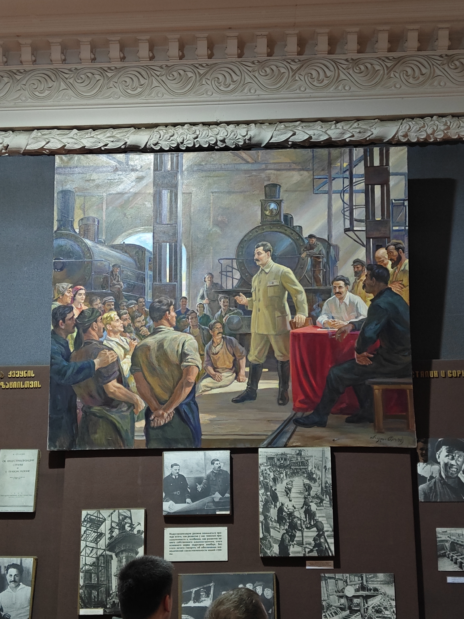 The image shows a large painting of Joseph Stalin addressing a crowd of people in what appears to be a factory setting with a train in the background. Stalin is wearing a light-colored uniform and stands in front of a red-draped table where another man is seated. A third man is sitting on a stool to the right of the table, facing away from the viewer. The crowd is a mix of men and women, all looking toward Stalin.
The painting is mounted on a wall, surrounded by other smaller pictures and documents. There are people in the foreground, likely museum visitors. The location is a museum, possibly located in the city of Unknown, Unknown. The artifacts surrounding the painting appear to be related to Stalin, and the industrialization period. The text on the documents is in Cyrillic.