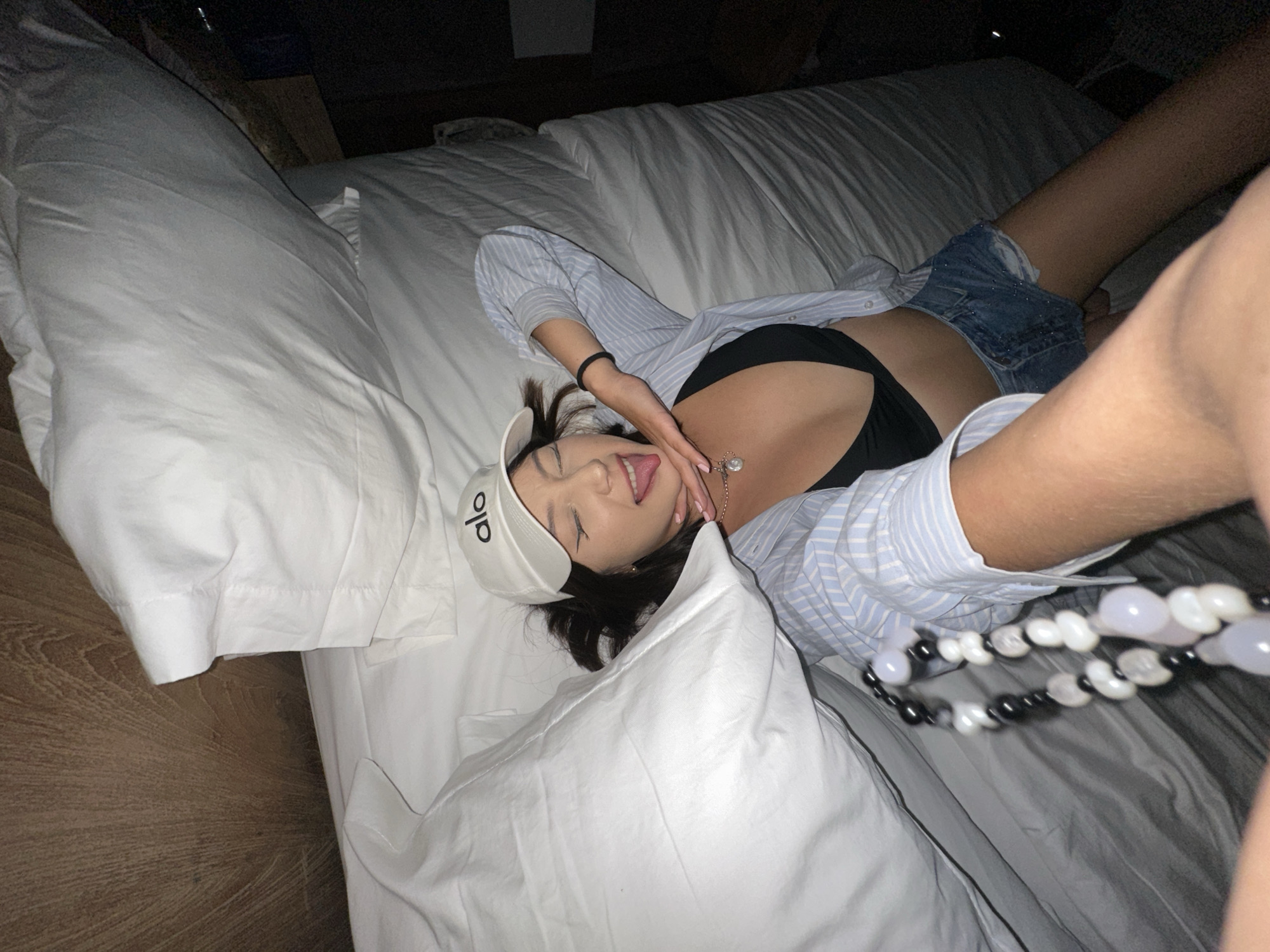 A woman is lying on a bed, wearing a white baseball cap, a blue and white striped shirt, a black bikini top, and denim shorts. She is sticking her tongue out and has her hand on her chin. She is also wearing a necklace with black and white beads. The bed is covered in white linens. The background appears to be a wooden headboard and a dark wall. The image is taken from a high angle, looking down at the woman. The location is specified as North Kuta, Indonesia.