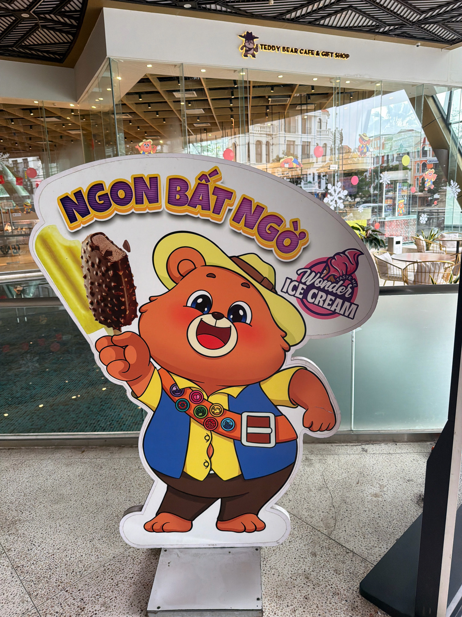 The image shows a cartoon bear mascot standing in front of a storefront with large glass windows. The bear is orange with big blue eyes and is wearing a yellow shirt, a blue vest, brown pants, and a straw hat. It is holding a chocolate-covered ice cream bar. A sign above the bear reads "NGON BẤT NGỜ" in large, colorful letters, which translates to "unbelievably delicious" in Vietnamese. To the right of the bear is a pink ice cream cone graphic with the text "Wonder ICE CREAM" and "TASTE ME." The storefront appears to be for a cafe and gift shop, as indicated by the sign "TEDDY BEAR CAFE & GIFT SHOP" visible above the entrance. The background shows the interior of the cafe, with tables, chairs, and displays. The flooring in front of the store is a speckled terrazzo. The scene is likely located in the city of Phú Quốc, Vietnam, given the Vietnamese text on the sign.