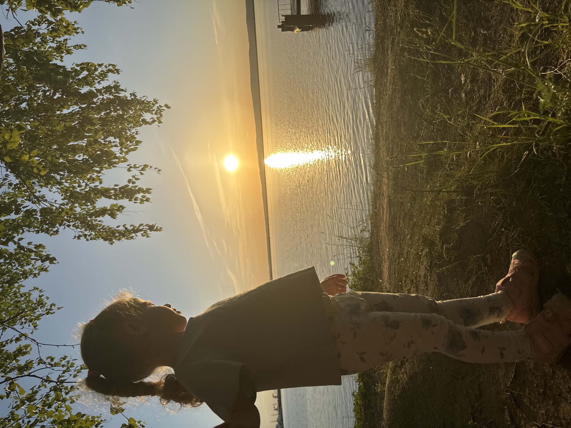 A child lies on the grass near a lake in Chaikovsky, Russia, at sunset. The sun is reflected on the water's surface. A tree is in the upper left corner of the image. A small dock extends into the lake. The child is wearing light colored pants and pink boots.