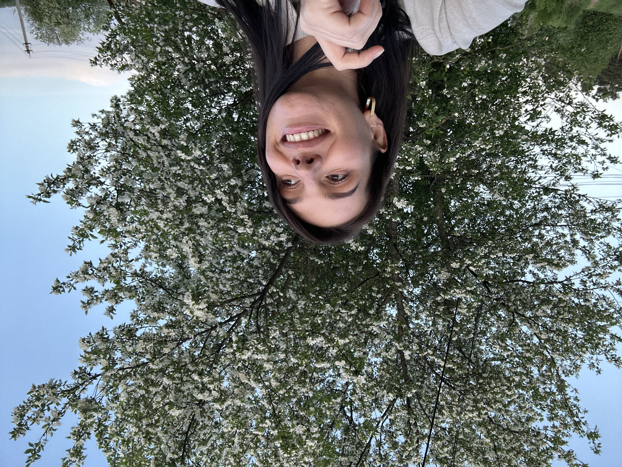 The image is an upside-down selfie of a woman with dark hair, light skin, and a gold hoop earring in her left ear. She is smiling and holding her hand near her face. Behind her is a tree in full bloom with small white flowers. In the background, there is a blue sky. Based on the location given, the scene likely depicts a woman taking a selfie with a blooming tree in Rusalevka, Russia.