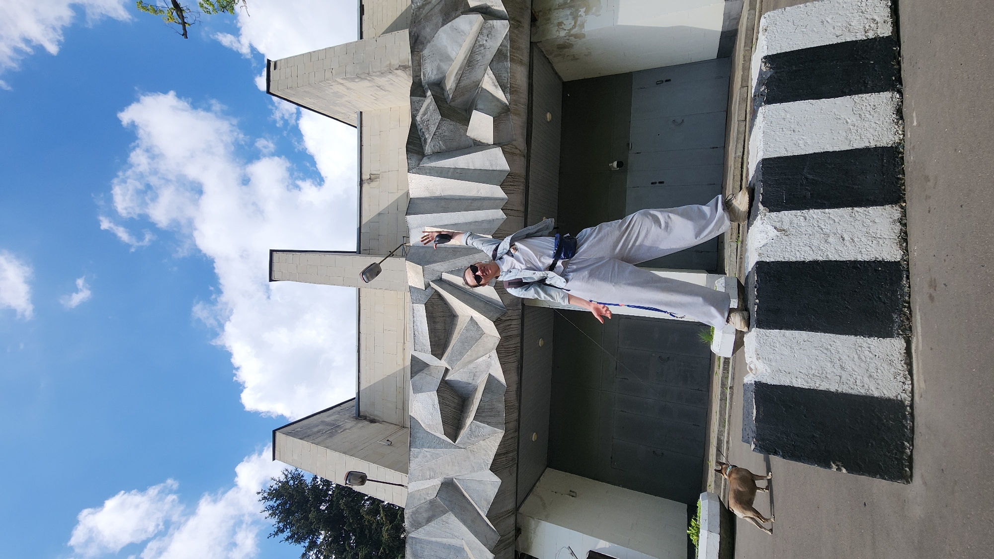 The image shows a person standing on a curb with alternating black and white stripes. The person is wearing light-colored pants, shoes, and a jacket. They are also wearing sunglasses. Behind the person is a building with a geometric facade. A small dog is visible at the bottom right corner of the image. The photograph was taken in Khimki, Russia.