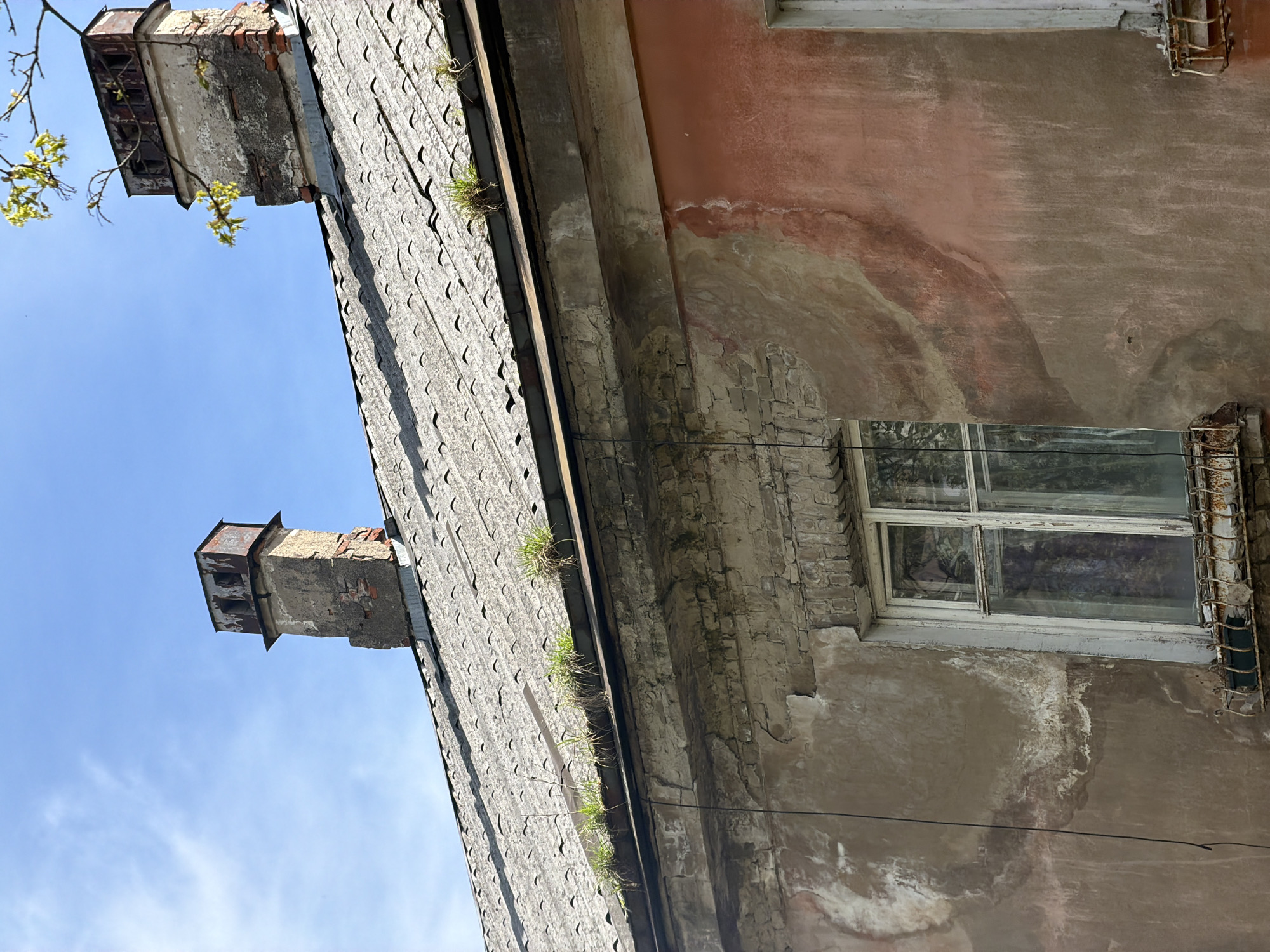 The image shows a section of an old, dilapidated building in Tokkari, Russia. The roof is made of gray, overlapping tiles with patches of green moss or grass growing along the edges. Two brick chimneys rise from the roof, one of which appears partially destroyed with exposed brick. The building's facade features a faded, peeling paint finish in shades of peach and gray. A single window with a white frame and a metal security cage is visible. The building appears to be in a state of disrepair, with crumbling brickwork and water stains on the walls. The sky is light blue with a few wispy clouds visible in the background. A small branch with green leaves is present at the upper left.