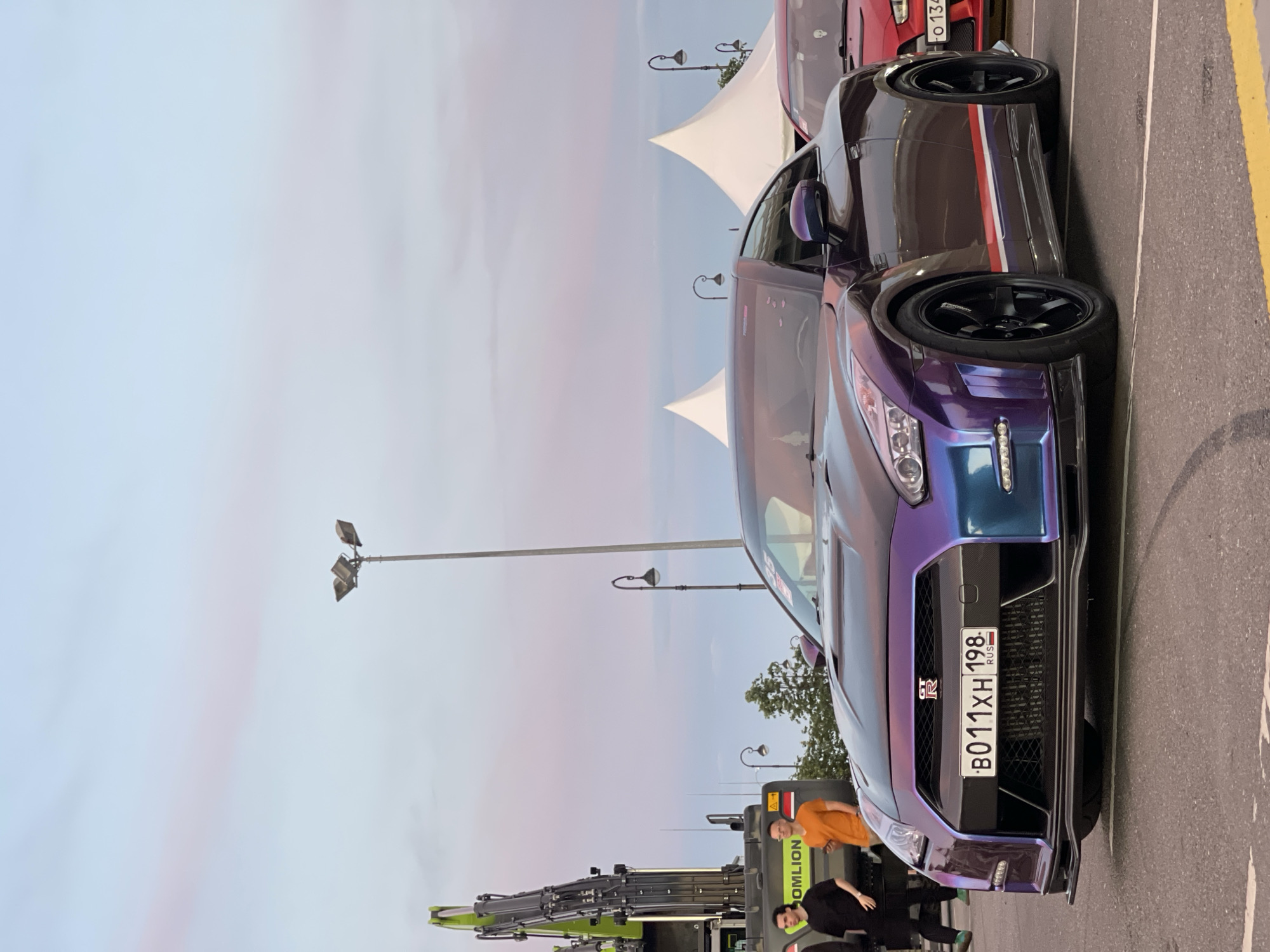 The image shows a purple and blue iridescent Nissan GT-R parked on asphalt. The car has a front license plate that reads "B011XH 198 RUS". To the left of the car, two men are standing near a large green construction vehicle. In the background, there are white tents, lampposts, and a cloudy sky with pink and blue hues. Another red car is partially visible to the right of the GT-R. The scene appears to be outdoors in an urban or event setting.
