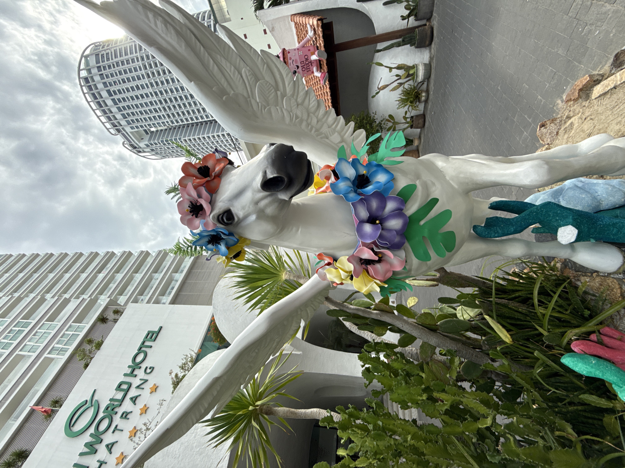 An exterior, low-angle, eye-level shot captures a white Pegasus statue adorned with a floral wreath around its neck and a crown of flowers on its head. The statue is positioned in front of a modern hotel building with multiple floors and balconies, and a distinctive curved building in the background. The hotel's signage reads "WORLD HOTEL NHA TRANG" with five gold stars. The foreground includes lush green cacti and other plants, as well as some colorful, abstract sculptures at the base of the Pegasus. A small, cartoon-like figure with a pink cup-shaped body and the words "OLD STUDIO" is perched on a tiled roof to the right of the statue. The sky is cloudy.