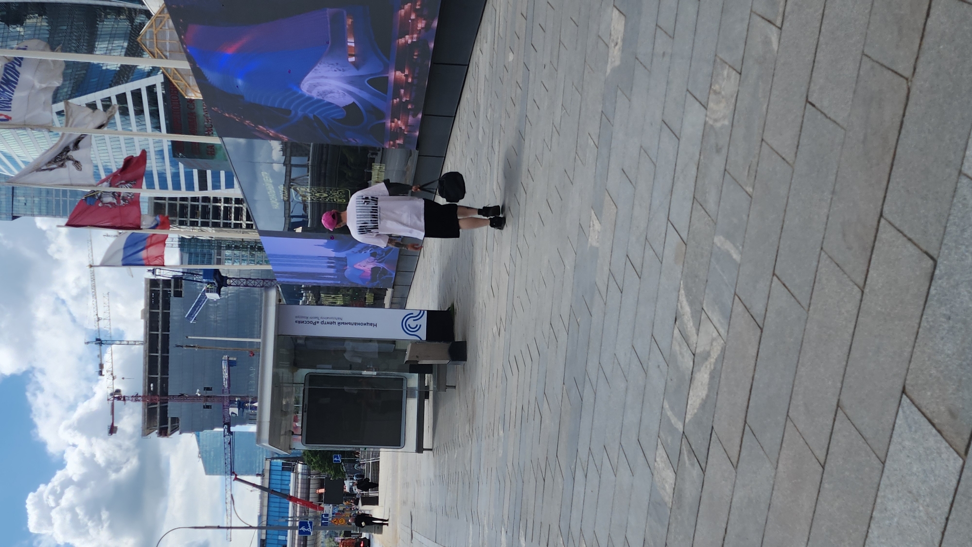 An outdoor shot shows a pedestrian walking away from the camera on a sidewalk made of gray pavers. The individual is wearing a pink baseball cap, a white t-shirt with black graphic print, black shorts, and black sneakers. They are carrying a black duffel bag in their right hand. Behind the pedestrian, there is a bus stop shelter with a digital display. Further in the background, a modern skyscraper construction site is visible, with cranes and partially built structures. Large digital billboards displaying colorful graphics are also present. The sky is partly cloudy.