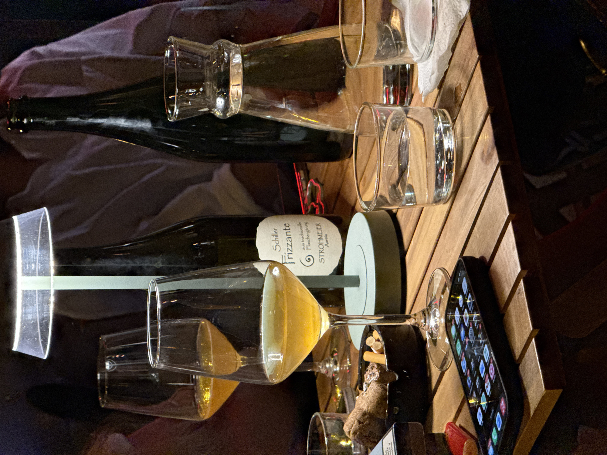 A wood-slatted table is topped with two bottles of wine, several glasses containing wine, and a smartphone. The lower bottle is labelled "Schiller Frizzante". An ashtray containing cigarette butts and a cork is also on the table. The photograph was likely taken indoors. The location, Vracar Urban Municipality, Serbia, cannot be confirmed solely from visual elements in the image. There are no people visible.