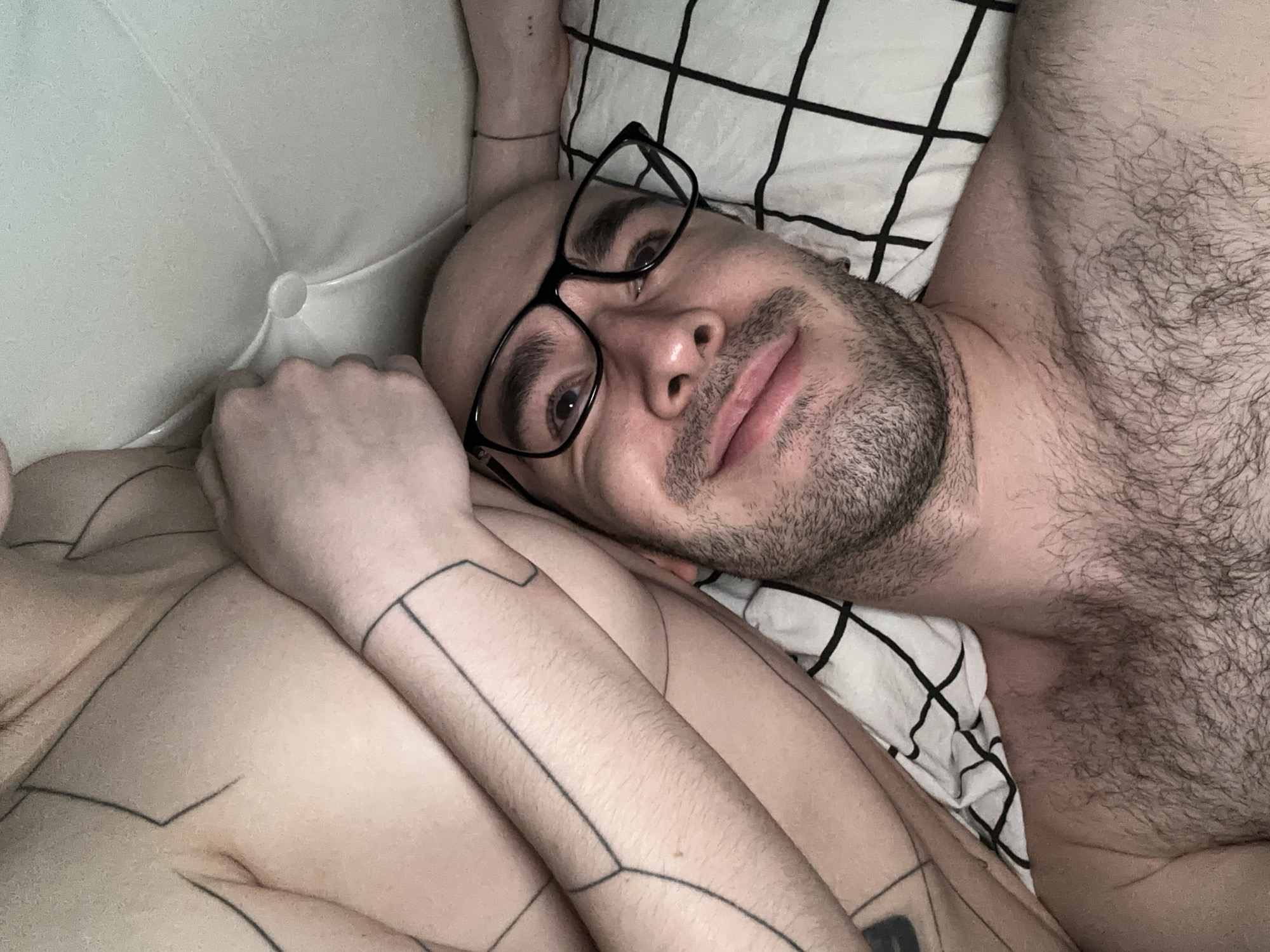 A close-up image shows a bald, light-skinned man with glasses lying in bed. He has a short beard and chest hair. He is on a white pillow with a black grid pattern. A person with geometric tattoos on their chest and arms is next to him. The location is not discernible from the image; Khimki, Russia cannot be determined.