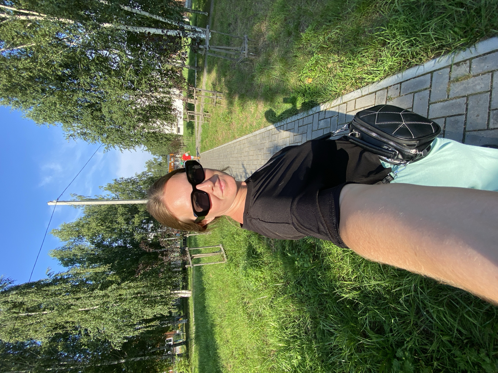 In Noviy, Russia, a selfie is taken by a woman standing on a paved walkway. She is wearing a black, short-sleeved, ribbed shirt, a light blue skirt, and black sunglasses. A black purse with white stitching is slung across her body. The walkway is bordered by a grassy area with several young trees, some supported by wooden frames. Further in the background, taller trees with white bark line the scene, and a utility pole with wires is visible. The sky is clear and blue, indicating a sunny day.