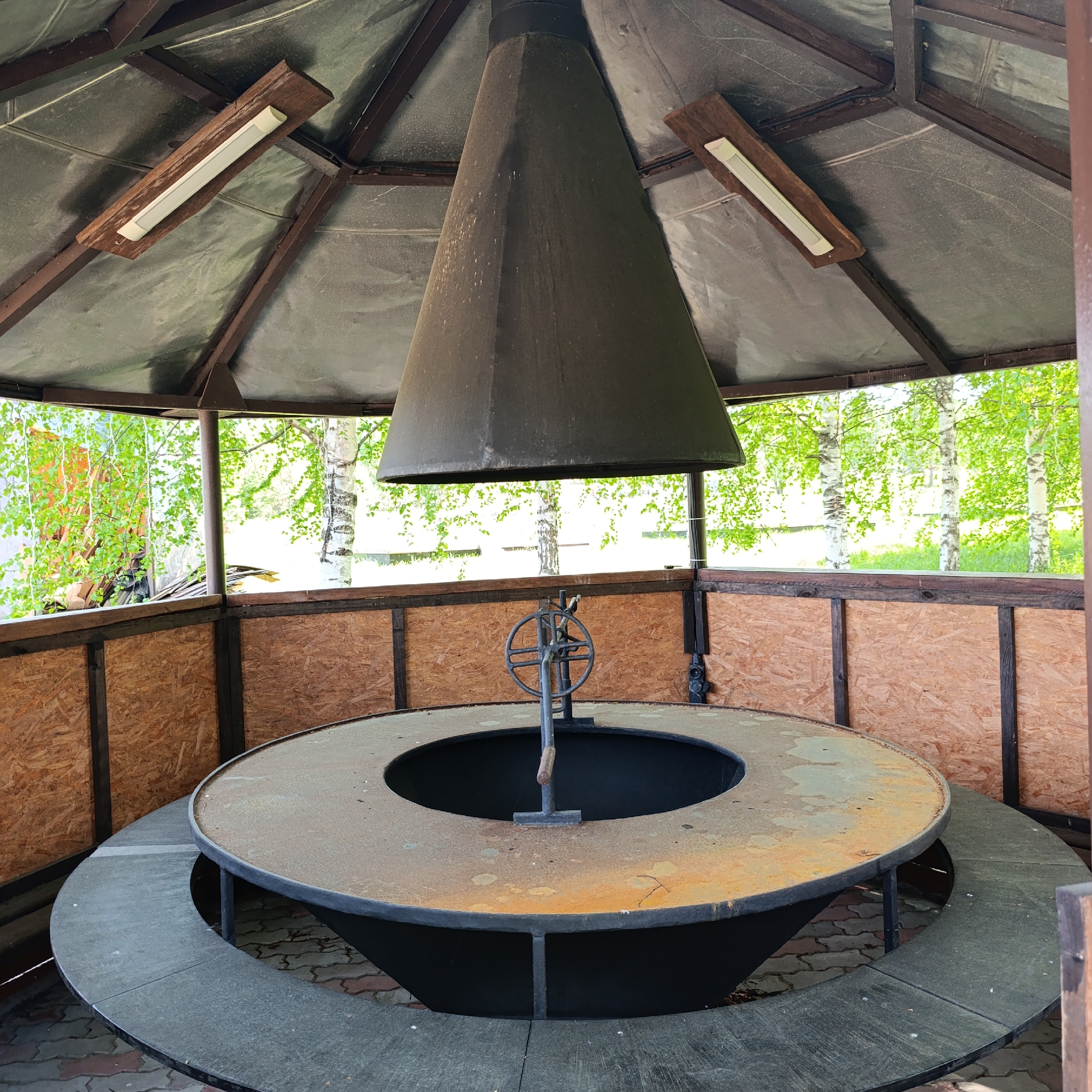 The image depicts a communal grill structure, possibly located in Prikamskii, Russia, though this is not explicitly confirmed by the image alone. The structure consists of a large, circular, waist-height grill with a central fire pit. A metal wheel mechanism hangs above the pit, presumably for adjusting the height of cooking implements. The grill is surrounded by a circular seating area at a lower level. A conical metal chimney hood hangs above the grill, presumably to exhaust smoke. The grill is housed within a hexagonal wooden gazebo structure with plywood paneling. Two fluorescent light fixtures are attached to the gazebo's ceiling. Outside the gazebo, trees and greenery are visible, suggesting an outdoor setting. The grill shows signs of wear and rust, indicating it has been used. There are no people visible in the image.