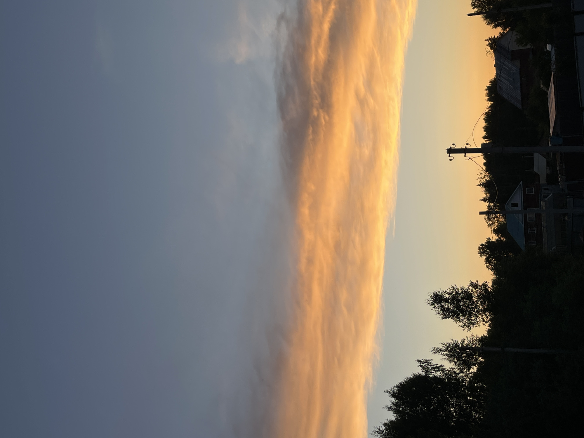 The image captures a serene sunset or sunrise scene over a rural or semi-rural landscape, possibly in Chaikovsky, Russia. The sky dominates the upper two-thirds of the frame, bisected by a prominent, elongated band of clouds. These clouds are brilliantly illuminated from below, glowing in vibrant orange and yellow tones, suggesting the golden hour of either dawn or dusk. Above this radiant cloud formation, the sky is a soft, hazy blue-grey, fading into lighter tones towards the right. Below the main cloud band, the sky transitions into warmer shades of yellow and orange as it meets the horizon.
The lower third of the image features a dark, silhouetted foreground. A dense line of trees, appearing as shadowy masses, stretches across the horizon. Among these trees, several buildings or houses are dimly visible as dark shapes, one with a hint of a red roof and lighter walls on the right side. A wooden utility pole with multiple crossarms and insulators stands distinctly on the right, rising above the tree line, with faint power lines extending from it. No people are visible, and no specific activity is taking place, contributing to the tranquil and atmospheric mood. There is no visible text in the image.