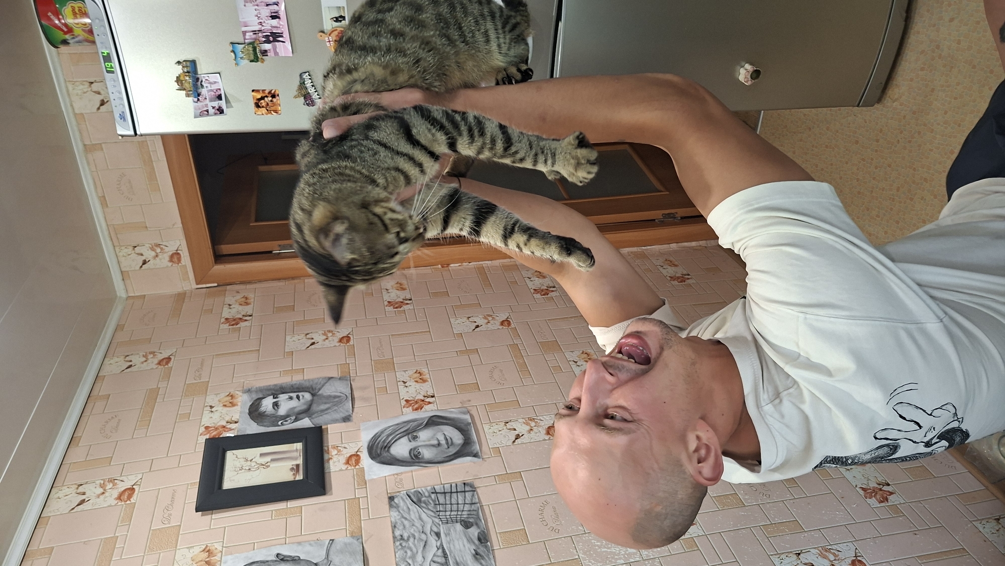 A bald man with an open mouth is holding a tabby cat above his head. The man is wearing a white t-shirt with a black drawing on the back. The cat is looking down, with its paws extended. In the background, there is a refrigerator with magnets on it, a door, and framed pictures on the wall. The room appears to be a kitchen. The wallpaper is a light pink with a floral pattern. This image was taken in the city of Myadzyel, Belarus.