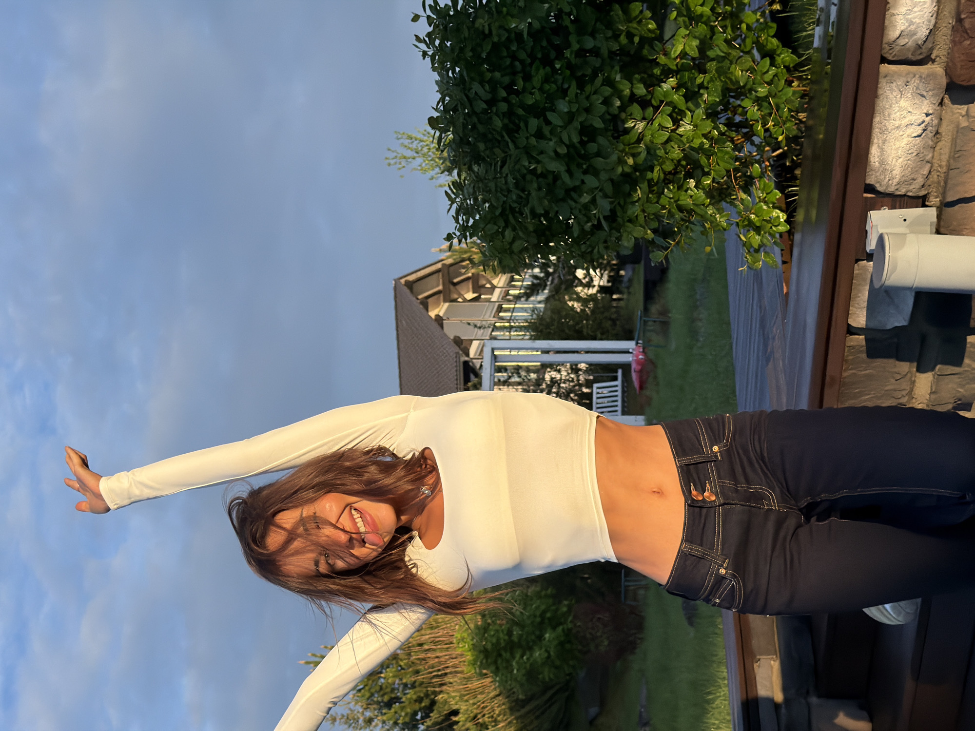 A young woman stands outside in Элита, Russia, smiling and extending her arms. She is wearing a white long-sleeved crop top and dark blue jeans. The background includes greenery, a house with a white porch, and a cloudy sky. Part of a structure made of wood and stone is visible on the right side of the frame.