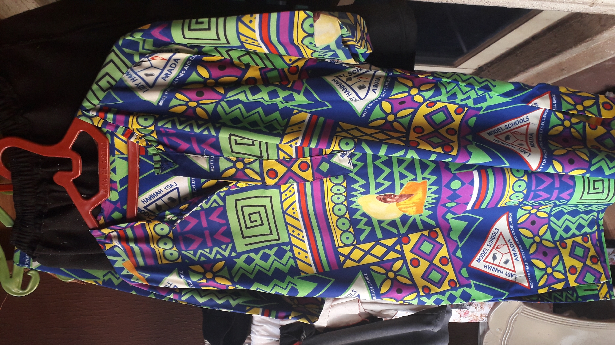 The image shows a garment, likely a uniform shirt or dress, hanging on a red plastic hanger. The garment has a complex, colorful pattern featuring geometric shapes like squares, triangles, and zigzags in shades of blue, green, yellow, purple, red, and white. Recurring motifs within the pattern include the text "LADY HANNAH" and "MODEL SCHOOLS AWADA" within a triangular logo. There is also text saying, "MOTTO: PACE SETTING AND EXCELLENCE." The specific location in the city of Unknown, Unknown, is unknown. Other items are visible in the background, including what appears to be a black garment, a window frame, a chair, and other indiscernible objects.