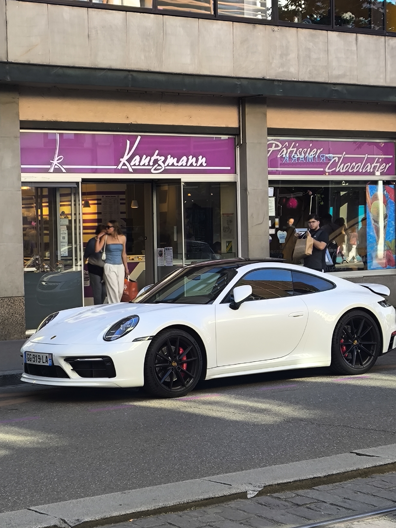 A white Porsche 911 is parked on a city street in front of a store with the signage "Kautzmann" and "Pâtissier Chocolatier." The store has a purple awning and glass doors. Two people stand in the entrance of Kautzmann, a woman in a blue top and white pants holding a bag. Another person is partially visible inside. At the entrance to "Pâtissier Chocolatier," a man in a black T-shirt exits carrying a brown paper bag. A tram track is visible in the street. The car has a license plate "GG-919-LA." The location is Strasbourg, France.