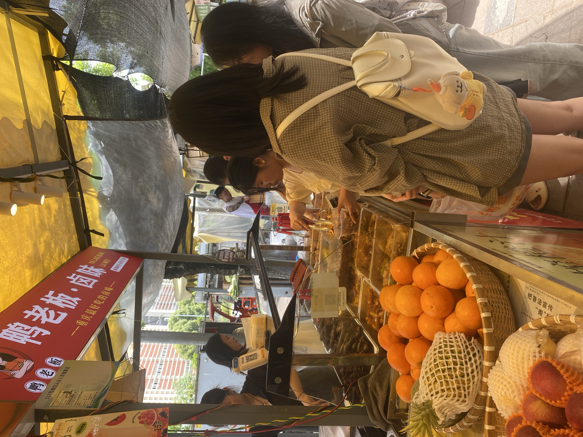 Here's a detailed description of the image:
**General Overview:**
The image depicts a street food stall, likely in Shapingba District, China, showcasing various prepared foods, fruits, and drinks. Several people are seen interacting with the stall, either browsing or purchasing items.
**Subjects and People:**
* Several young women are looking at the food selection at the stall. One wears a plaid jacket with a white backpack, while another has a gray top.
* An individual, possibly the vendor, is behind the counter.
* Other people are visible in the background, suggesting a busy street environment.
**Objects:**
* The food stall has a red sign with Chinese characters. The sign reads "鸭老板・卤味" (Ya Lao Ban・Lu Wei), suggesting the stall specializes in duck and braised dishes.
* A variety of food items are displayed in containers, including braised items, fruits, and possibly drinks.
* Baskets are filled with oranges and other fruits, such as pears and lychees.
**Actions and Interactions:**
* The women are looking at and possibly selecting food items from the stall.
* Money may be changing hands between the vendor and customers.
**Environment:**
* The food stall is set up outdoors on a paved street or sidewalk.
* Buildings and greenery can be seen in the background.
* Overhead, there's a mesh covering providing shade.