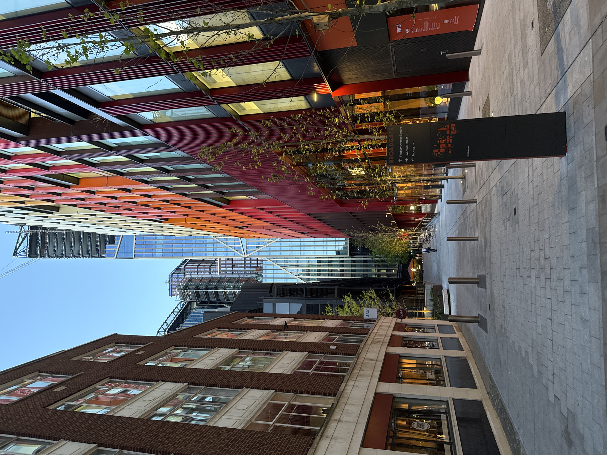 The image shows a street view in London, United Kingdom, featuring a mix of modern and traditional architecture. On the left, a tall building with a vibrant, multi-colored facade (ranging from orange to red) and reflective glass windows rises into a clear, blue sky. Adjacent to it, construction of other skyscrapers is visible.
On the right, a brick building with stone detailing is situated, possibly containing retail spaces at ground level. The street is paved with gray stone blocks and lined with metal bollards. A black sign indicates directions to "Broadgate," "Finsbury Avenue Square," "Liverpool Street," and "Moorgate." A Costa coffee shop is also visible. Above, trees with sparse foliage are present. The scene appears to be taken during daylight hours.