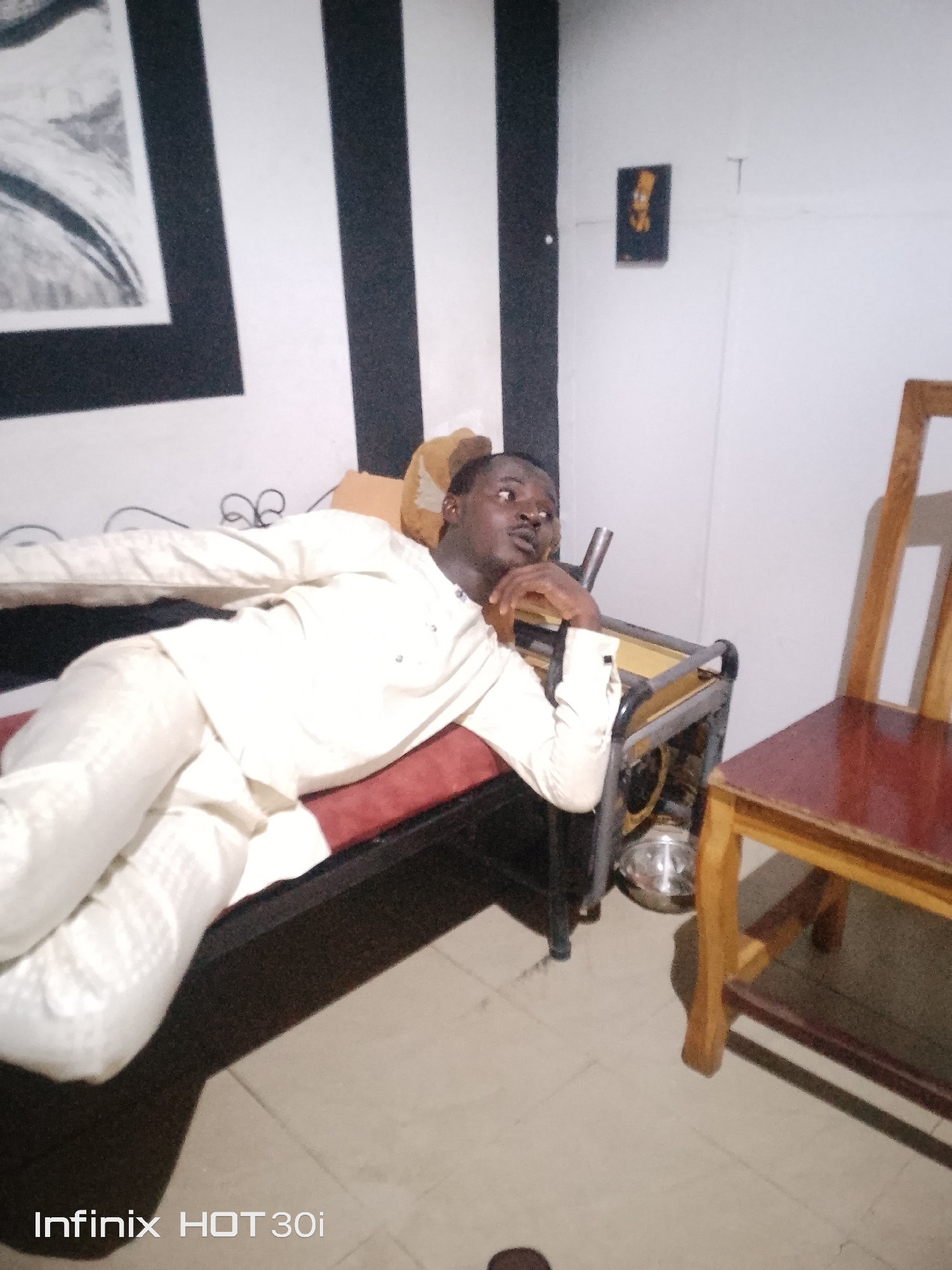 A man in a cream-colored outfit lies on a red chaise lounge. The lounge is positioned in front of a wall with vertical black and white stripes. To the right of the lounge, there is a wooden chair with a dark red seat. A small framed picture, possibly of a cartoon character, is hanging on the wall behind the man. The floor is tiled with a light beige color. The overall lighting is dim. The scene appears to be indoors, in what could be a room in a residential or commercial setting.