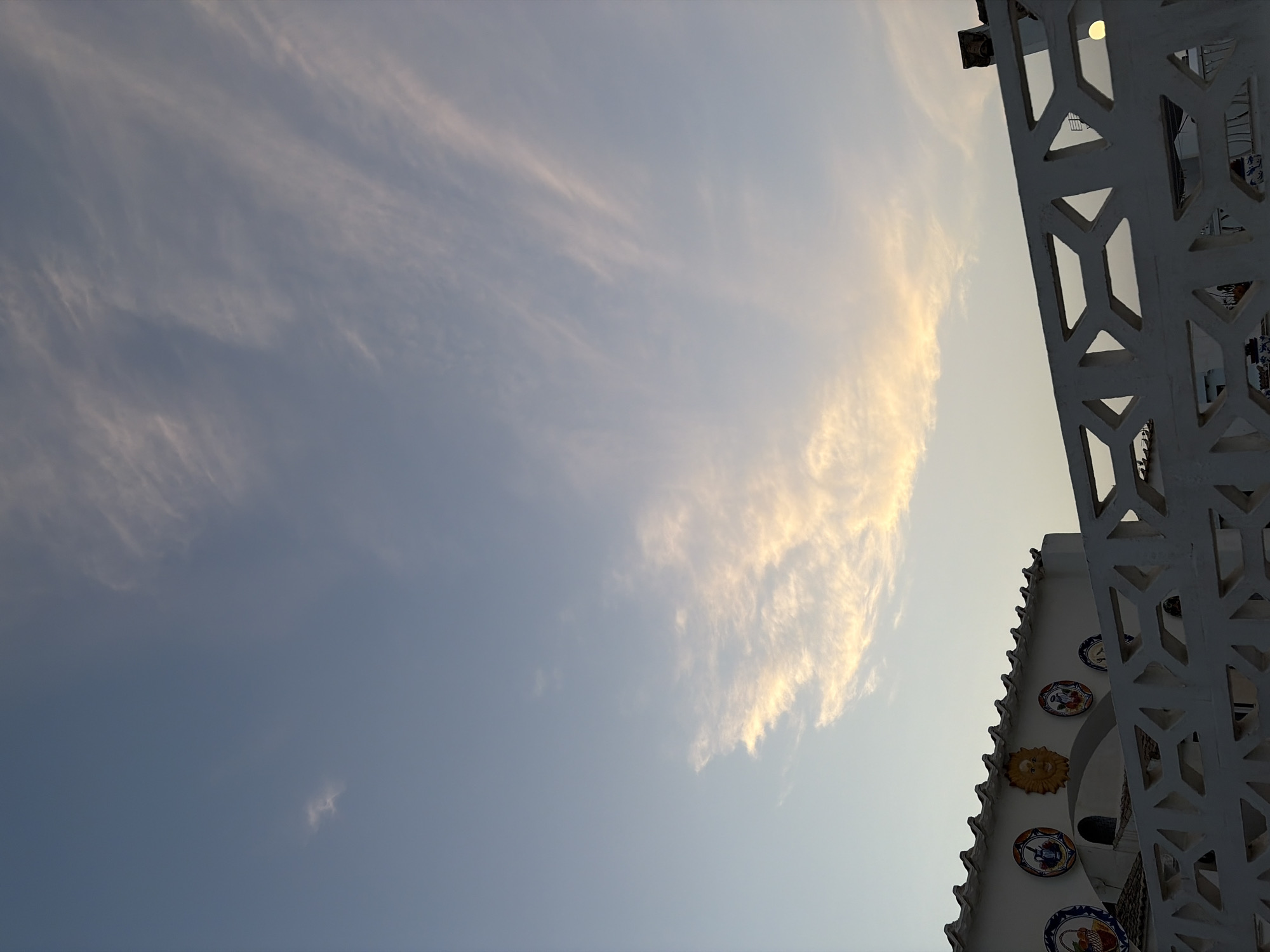 The image captures a late afternoon or early evening sky over Torrevieja, Spain, characterized by a pale blue expanse with streaks of wispy, orange-tinged clouds. In the foreground, a white, decorative lattice fence runs horizontally across the lower portion of the frame. Behind the fence, a white building with a tiled, zig-zagging roofline is visible. The building is adorned with circular ceramic tiles featuring various designs, including a sun face and depictions of baskets of fruit and pitchers. The overall atmosphere is serene and suggestive of a coastal setting. There are no people or active interactions depicted in the image.