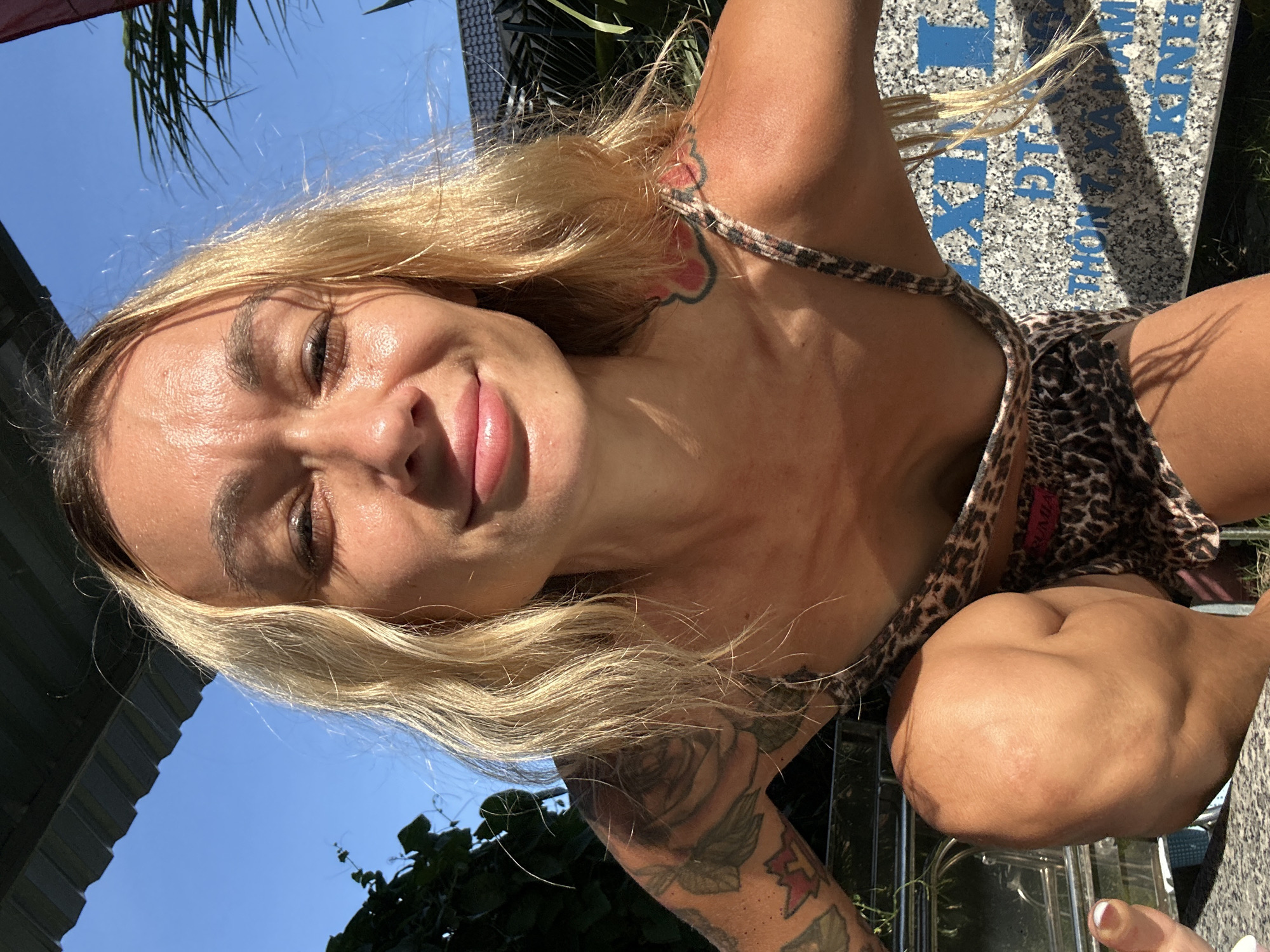 A blonde woman with tattoos on her arms and chest is photographed outdoors in Mũi Né, Vietnam. She is wearing a leopard-print top and shorts. The woman is crouching, looking up at the camera. The background shows a blue sky, green foliage, and a building with a tiled roof. There are also some blue and white signs in the background. The lighting suggests it is daytime, likely sunny.