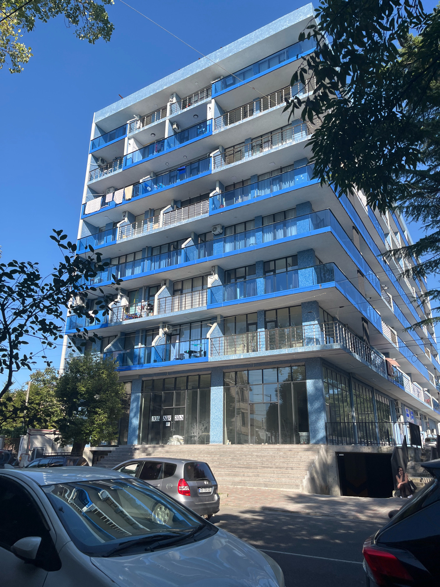 The image shows a multi-story building with a bright blue and white facade, located on a sunny day in Kobuleti, Georgia. The building features numerous balconies with blue glass railings and white trim. Laundry is visible drying on some of the balconies. At the base of the building, there are large glass windows and doors, with a set of wide concrete stairs leading up to the entrance. A dark entrance to what appears to be a parking garage is situated to the right of the stairs. Several cars are parked on the street in front of the building. A woman is seated on the concrete stairs near the parking garage entrance, looking to the right. Trees with green leaves partially obscure the building. The sky is clear and blue.
