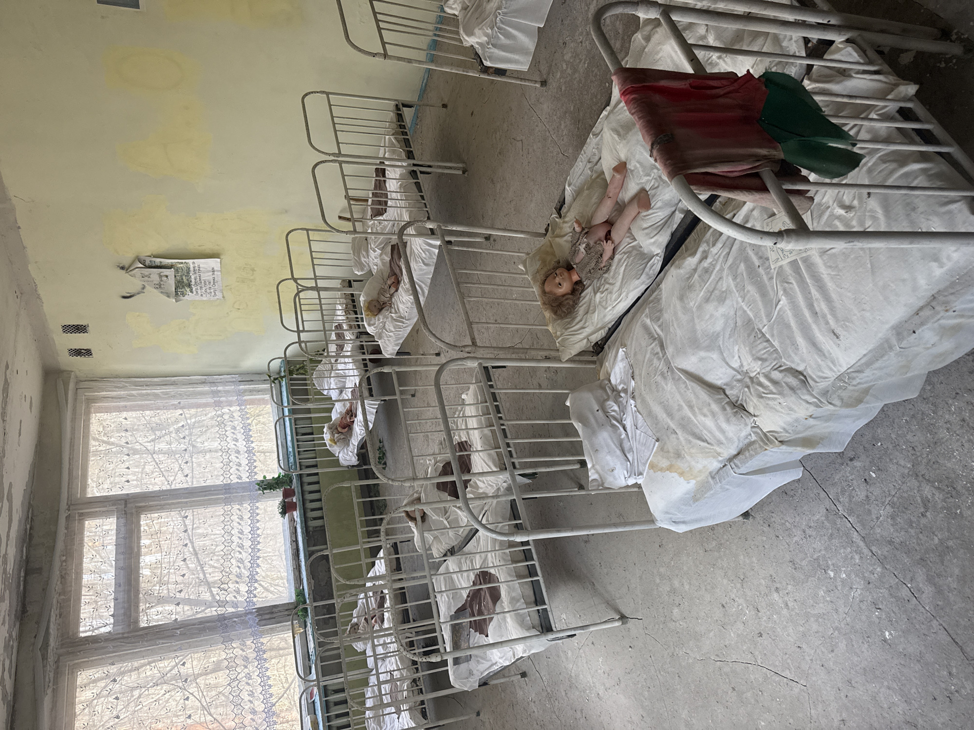 The scene is an abandoned room, likely a nursery or orphanage, in the city of Prypiat, Ukraine. Rows of small, metal-framed beds are lined up, each covered with stained white sheets. Several dolls lie in the beds. The room shows signs of disrepair: peeling yellow paint on the walls, a tattered poster. A window to the left reveals a view of bare trees. The floor is cracked concrete. A piece of red and green cloth sits on one of the beds.