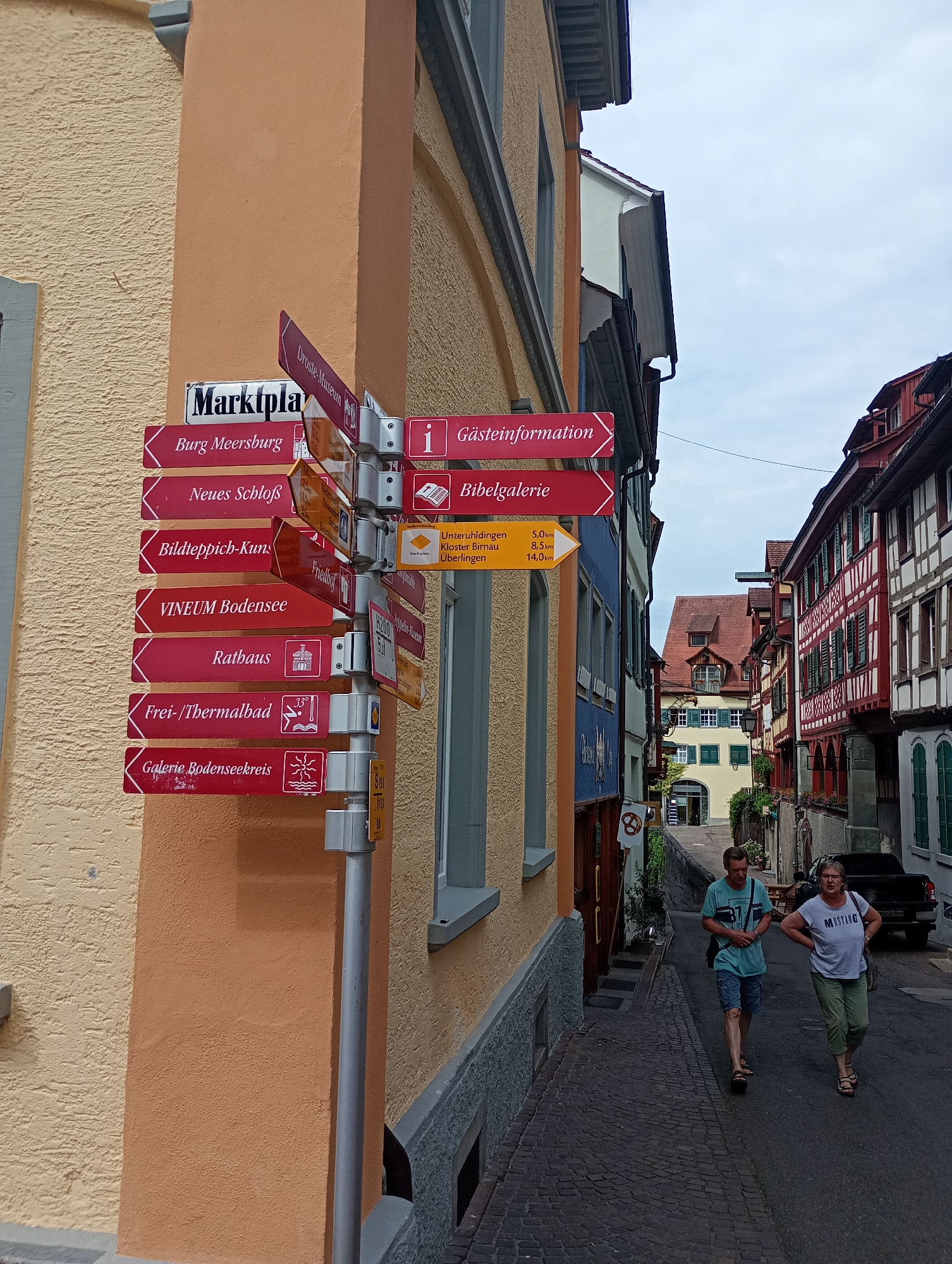 The image shows a narrow cobblestone street in Meersburg, Germany. On the left, a building with a textured orange facade is visible. A tall, silver metal pole stands on the right side of the street, adorned with multiple directional signs. These signs point to various locations, including "Marktplatz," "Burg Meersburg," "Neues Schloß," "Bildteppich-Kunst," "Vineum Bodensee," "Rathaus," "Frei-/Thermalbad," and "Galerie Bodenseekreis." Some signs also include distance markers in kilometers. On the right side of the street, there are half-timbered buildings with red and white facades. Two people are walking up the street, a man and a woman. The man is wearing a teal t-shirt and blue shorts, and the woman is wearing a white t-shirt with "Mustang" printed on it and green pants. The sky is overcast.