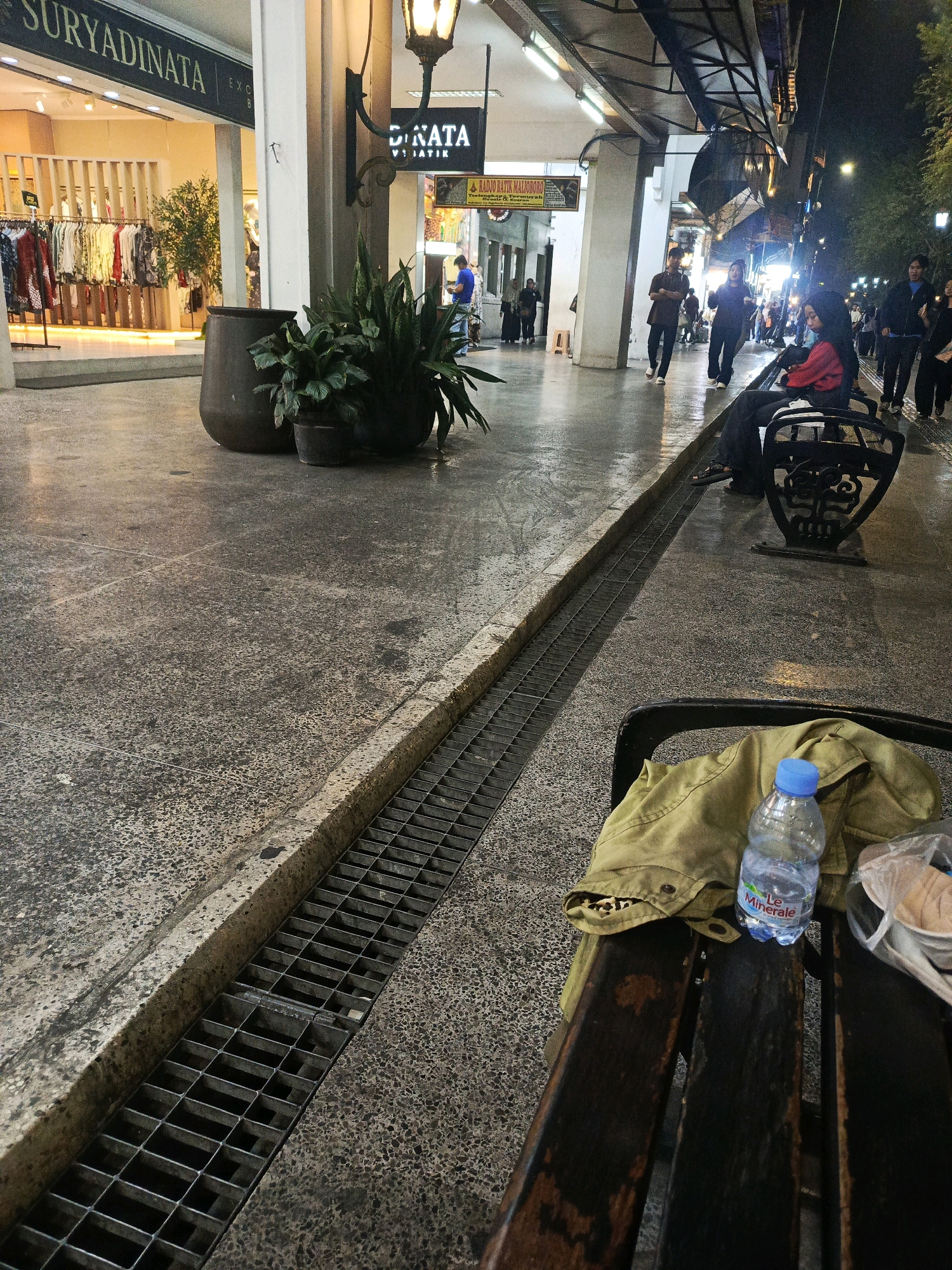 The image depicts a street scene at night in Malioboro Street, Yogyakarta, Indonesia. A concrete pathway, damp with what appears to be recent rain, is in the foreground. A metal grate runs along the side of the pathway, channeling water. In the lower right, a dark wooden bench is visible with a tan jacket draped over it, alongside a partially filled plastic bottle of "Le Minerale" water and a white plastic bag containing food items.
Further into the scene, people are walking and standing. A woman in a red top and headscarf is seated on a black metal bench. Several individuals are walking along the pathway, some silhouetted against bright shop lights. On the left side of the street, shop fronts are illuminated, including a store with "SURYADINATA" visible on its awning and clothing displayed on racks. A large, dark ceramic pot with plants sits near the shops, along with a smaller potted plant. The overall lighting suggests it is nighttime, with streetlights and shop lights providing illumination.