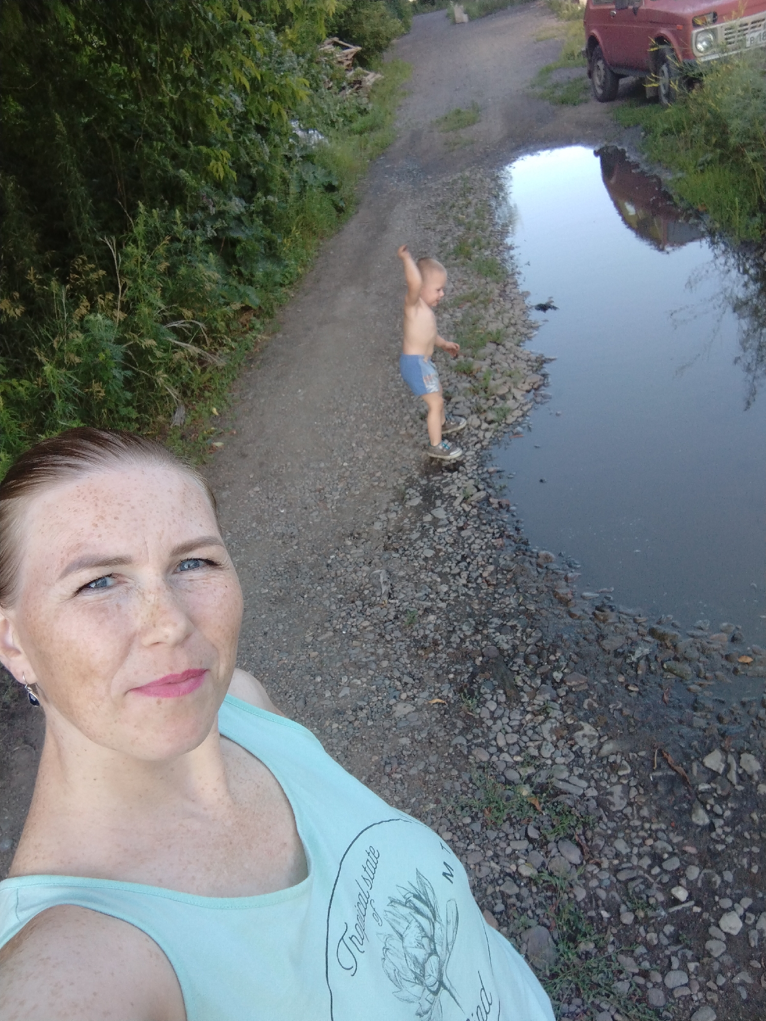 In the city of Komsomolsk-on-Amur, Russia, a woman with fair skin and freckles, wearing a light blue tank top, takes a selfie. Her hair is pulled back, and she is looking towards the camera with a slight smile. To her left, a young boy, wearing blue shorts and sneakers, is standing on a gravel path near a large puddle of water, his arm raised. A red car is parked further up the path, its reflection visible in the puddle. The surrounding area is characterized by a gravel road, lush green foliage, and a small body of water.