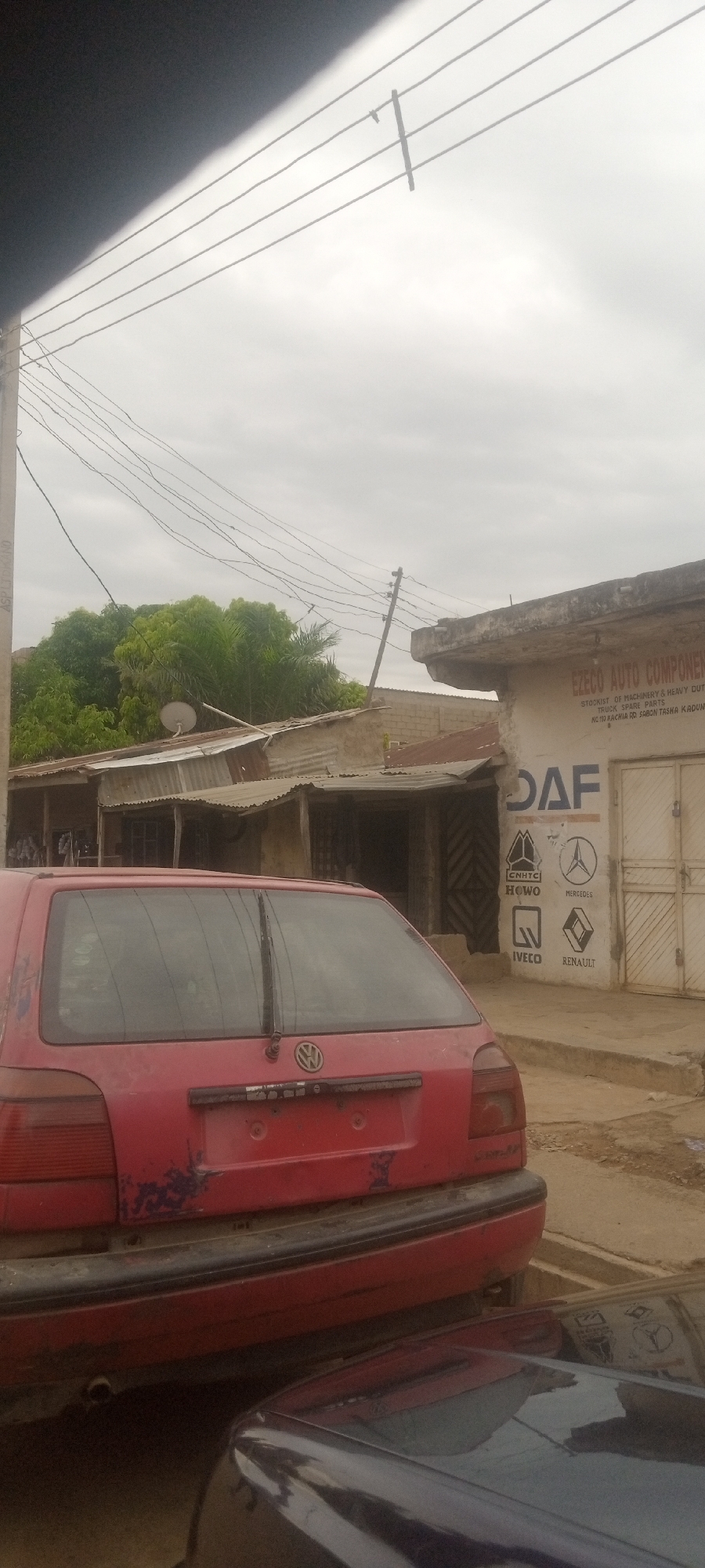 The image captures a street scene in Sabon Tasha, Nigeria. A red Volkswagen Golf stands in the foreground. Adjacent to the Golf, a building displaying the sign "EZECO AUTO COMPONENTS" advertises truck spare parts and lists brands such as DAF, CNHTC HOWO, IVECO, Mercedes, and Renault. The area appears to be a commercial or industrial zone, with other buildings visible in the background, some having corrugated iron roofs. Power lines crisscross the sky. The weather is overcast.